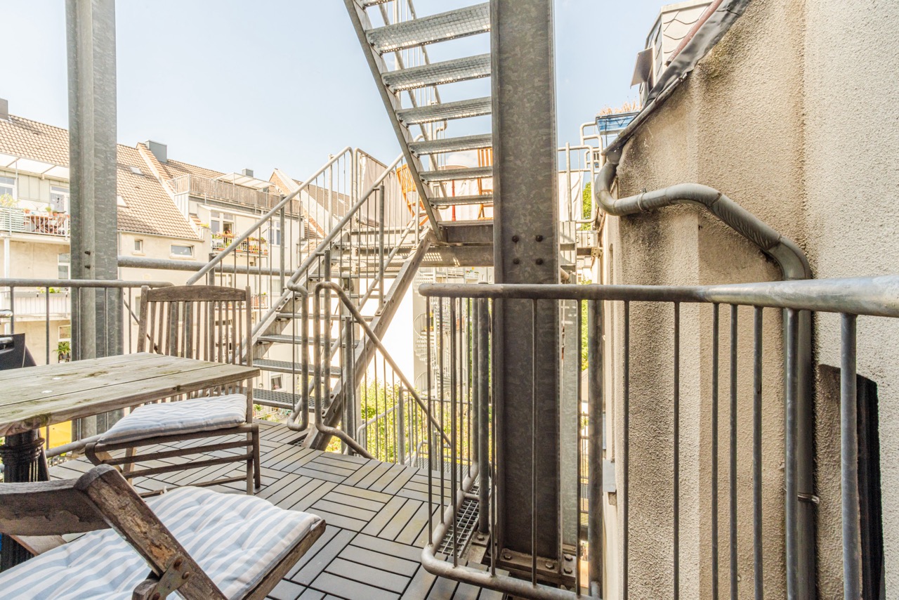 Balkon mit Holztisch, zwei Stühlen und Metalltreppe im Freien, umliegende Wohngebäude sichtbar.