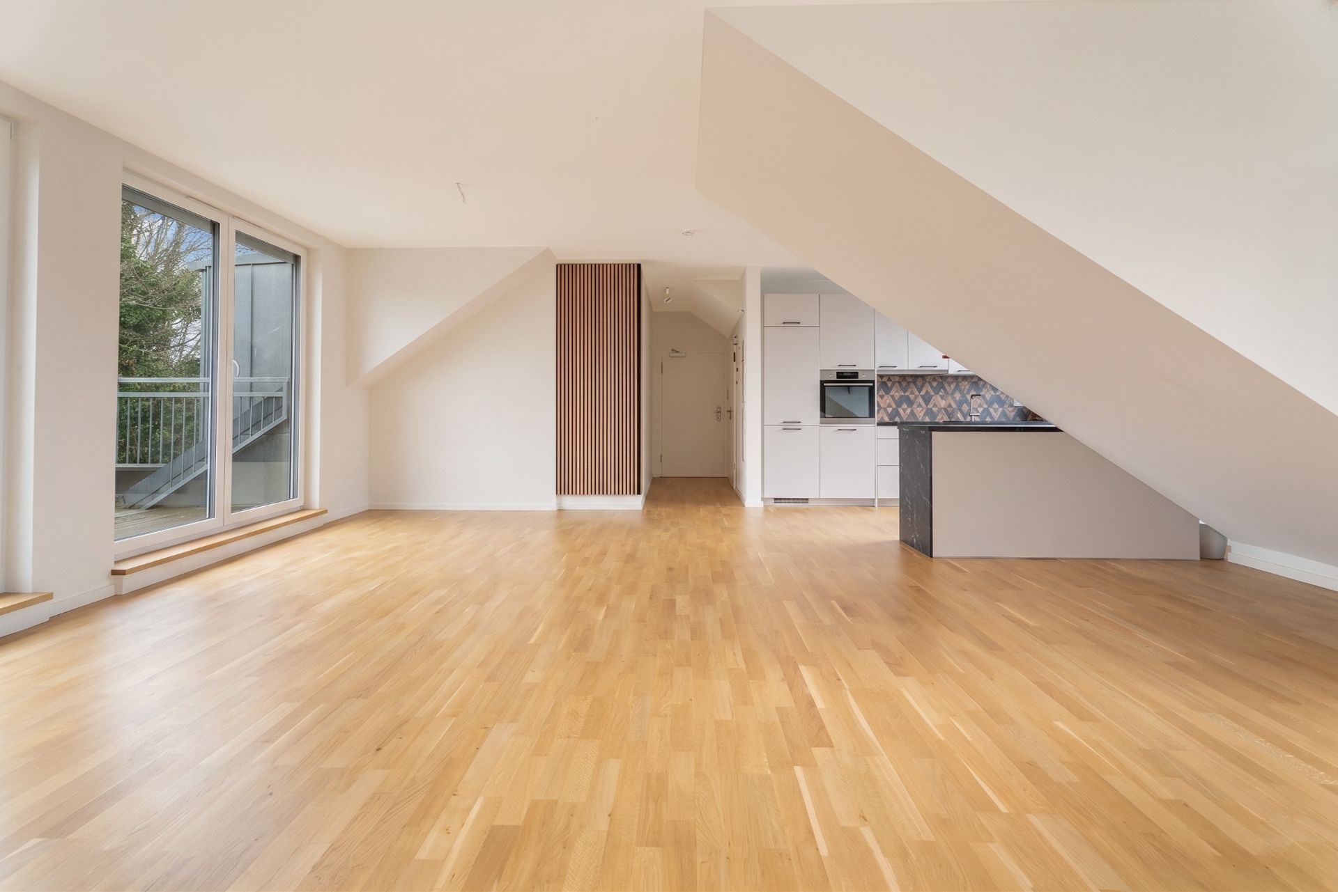 Heller Wohnraum mit Parkettboden, großer Fensterfront und offener Einbauküche im Dachgeschoss.