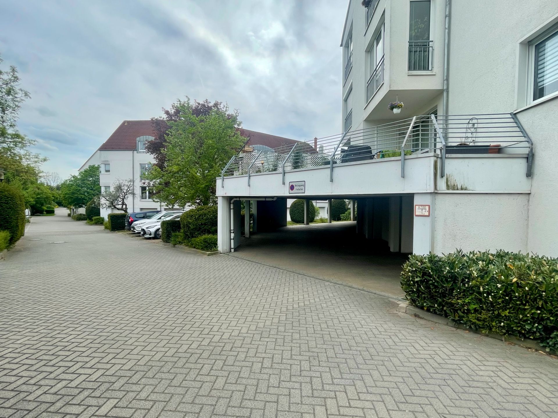 Wohngebäude mit offenen Parkplätzen und gepflasterter Zufahrt, Bäume und Sträucher am Rand.