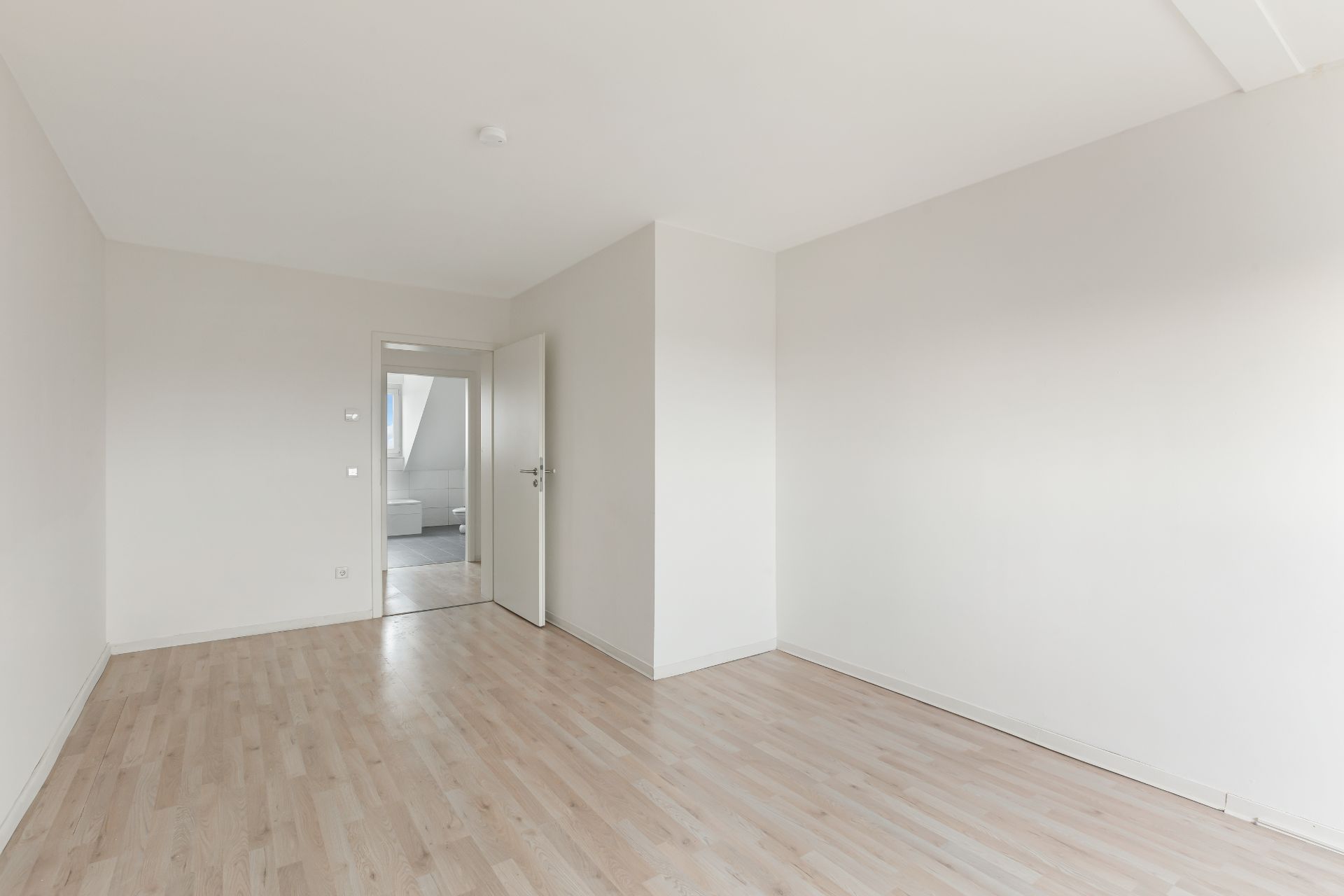 Leerer, heller Raum mit Holzboden und weißer Wand, Tür führt in einen weiteren Raum.