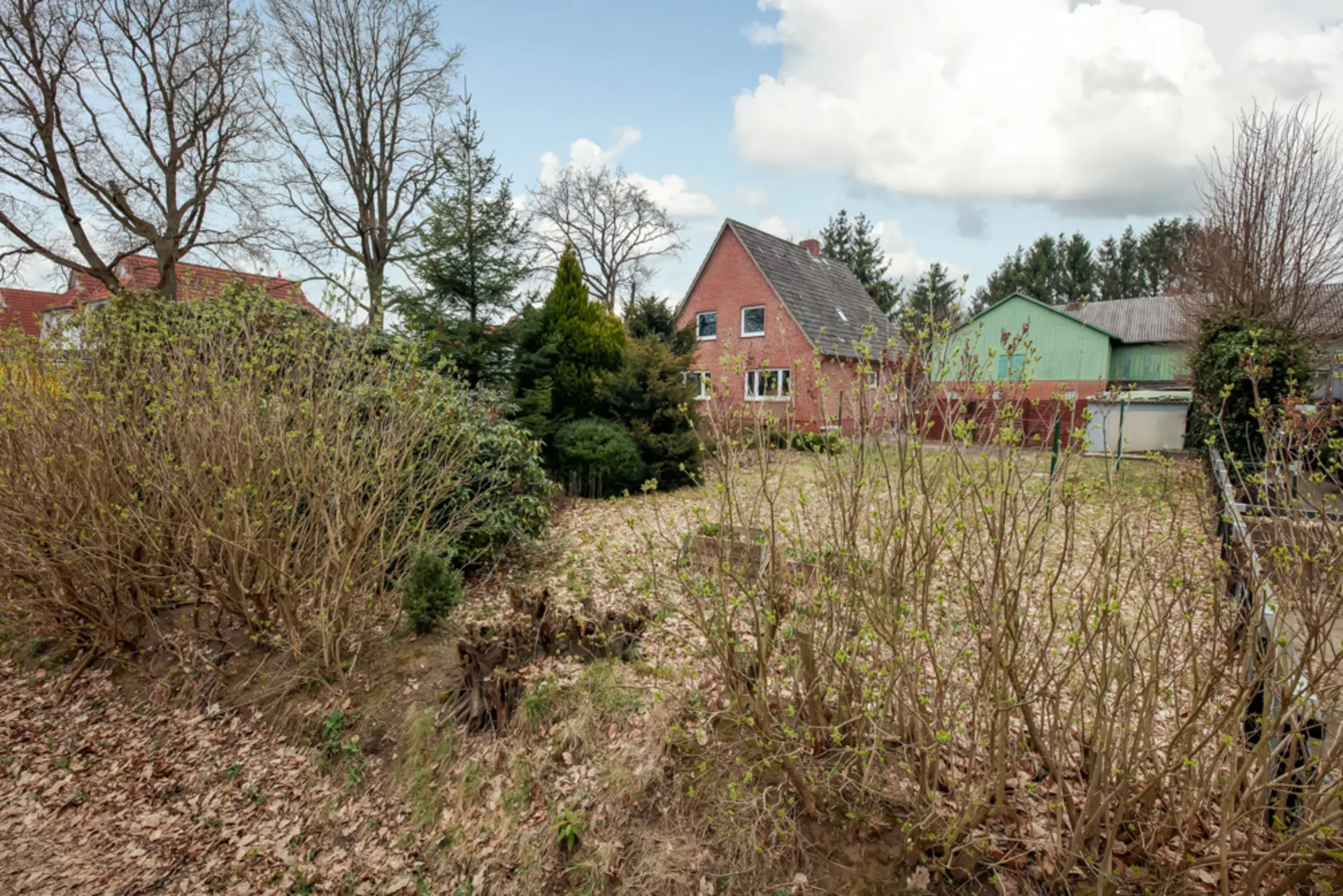 Garten mit Sträuchern und Bäumen, im Hintergrund ein rotes Wohnhaus und eine grüne Scheune.