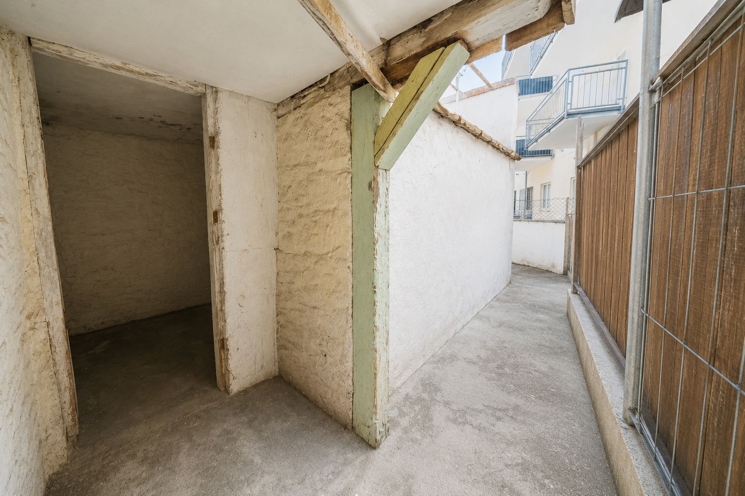 Überdachter, schmaler Durchgang mit Zugang zu einem Abstellraum, Wände und Boden sind aus Beton.