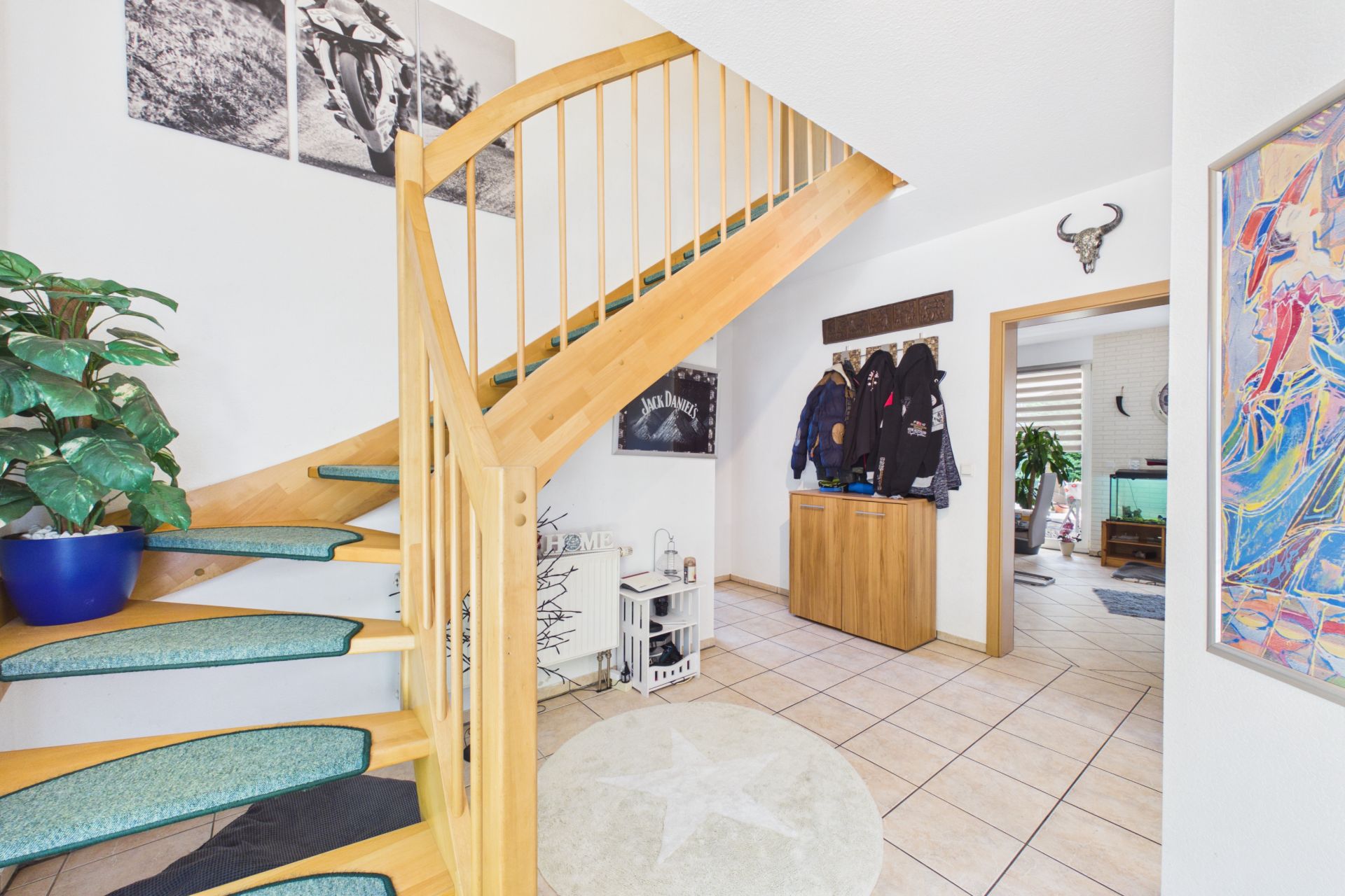 Heller Flur mit Holztreppe, runder Teppich, Pflanzen und Garderobe mit Jacken.