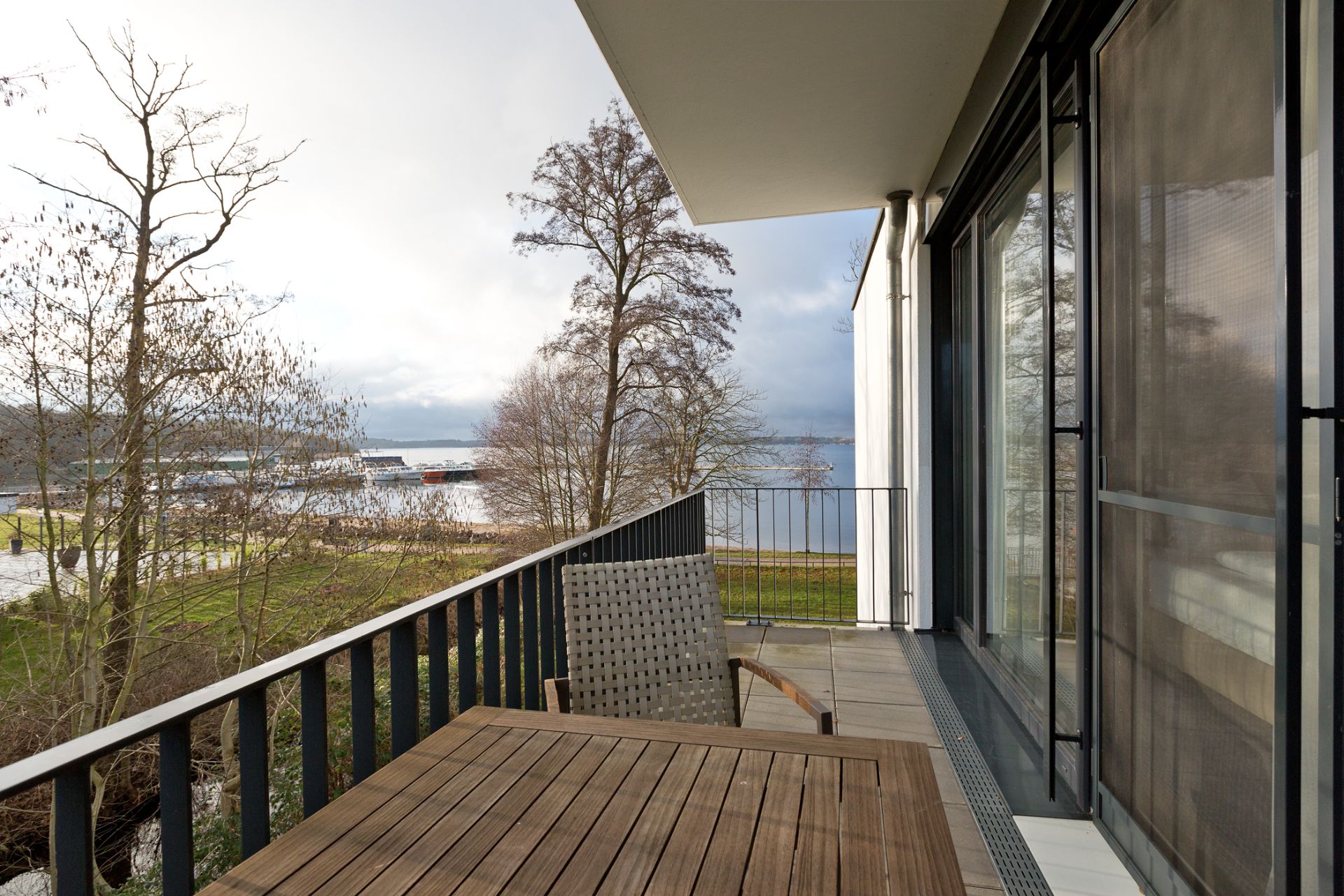 Balkon mit Holztisch, Stuhl und Ausblick auf Bäume, Wiese und Wasser.