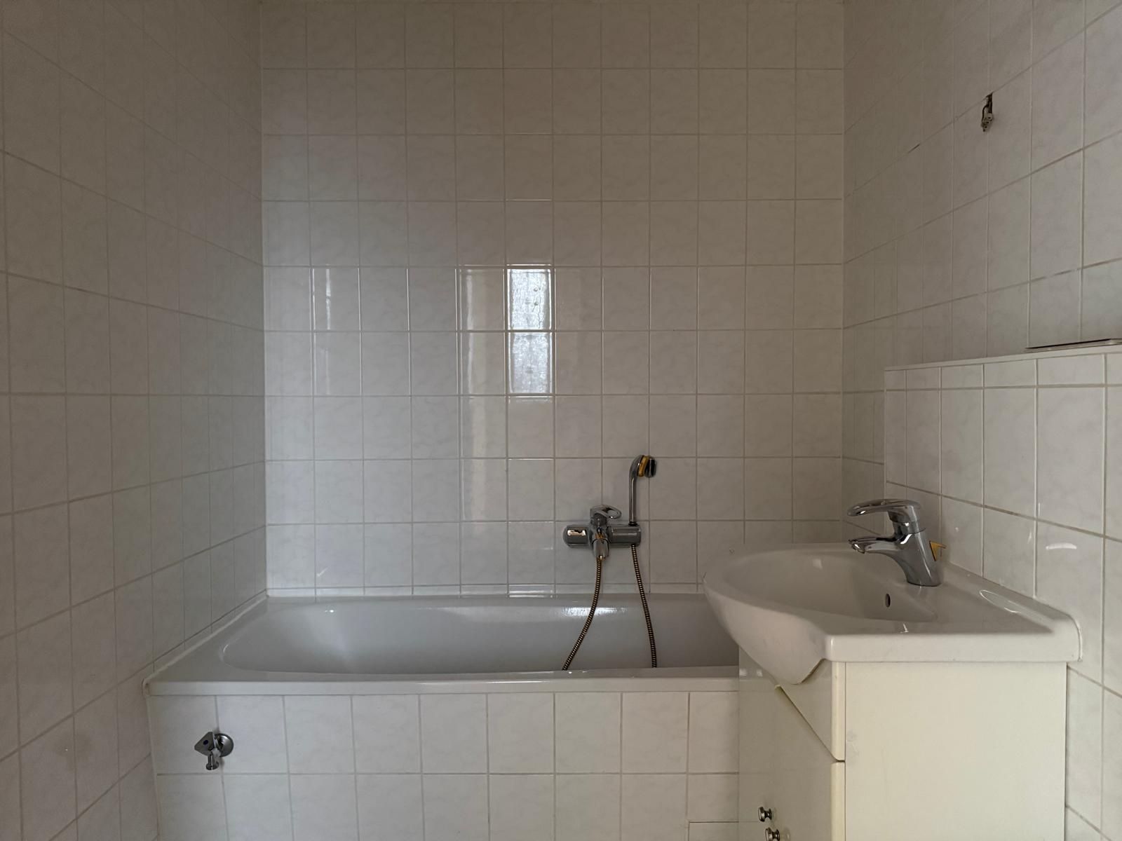 Bad mit weiß gefliester Badewanne und Waschbecken, Wandarmatur zwischen beiden.