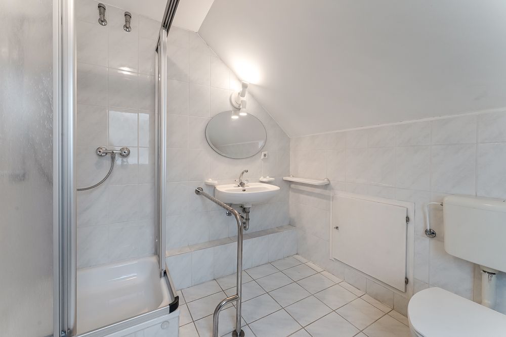 Helles Badezimmer mit Dusche, Waschbecken, rundem Spiegel und Toilette unter Dachschräge.