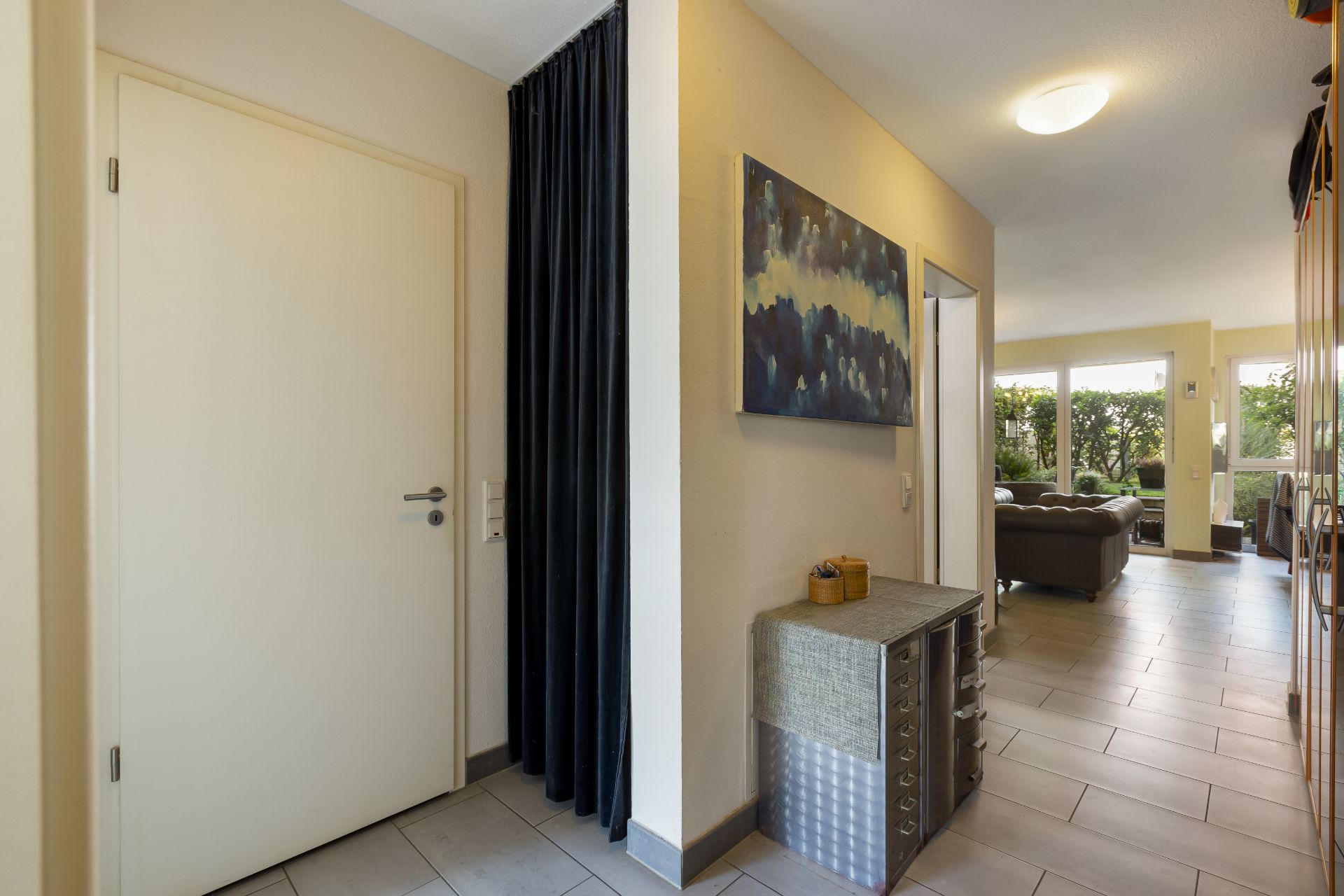 Heller Flur mit Kommode, Wandbild und Blick ins Wohnzimmer mit großen Fenstern.