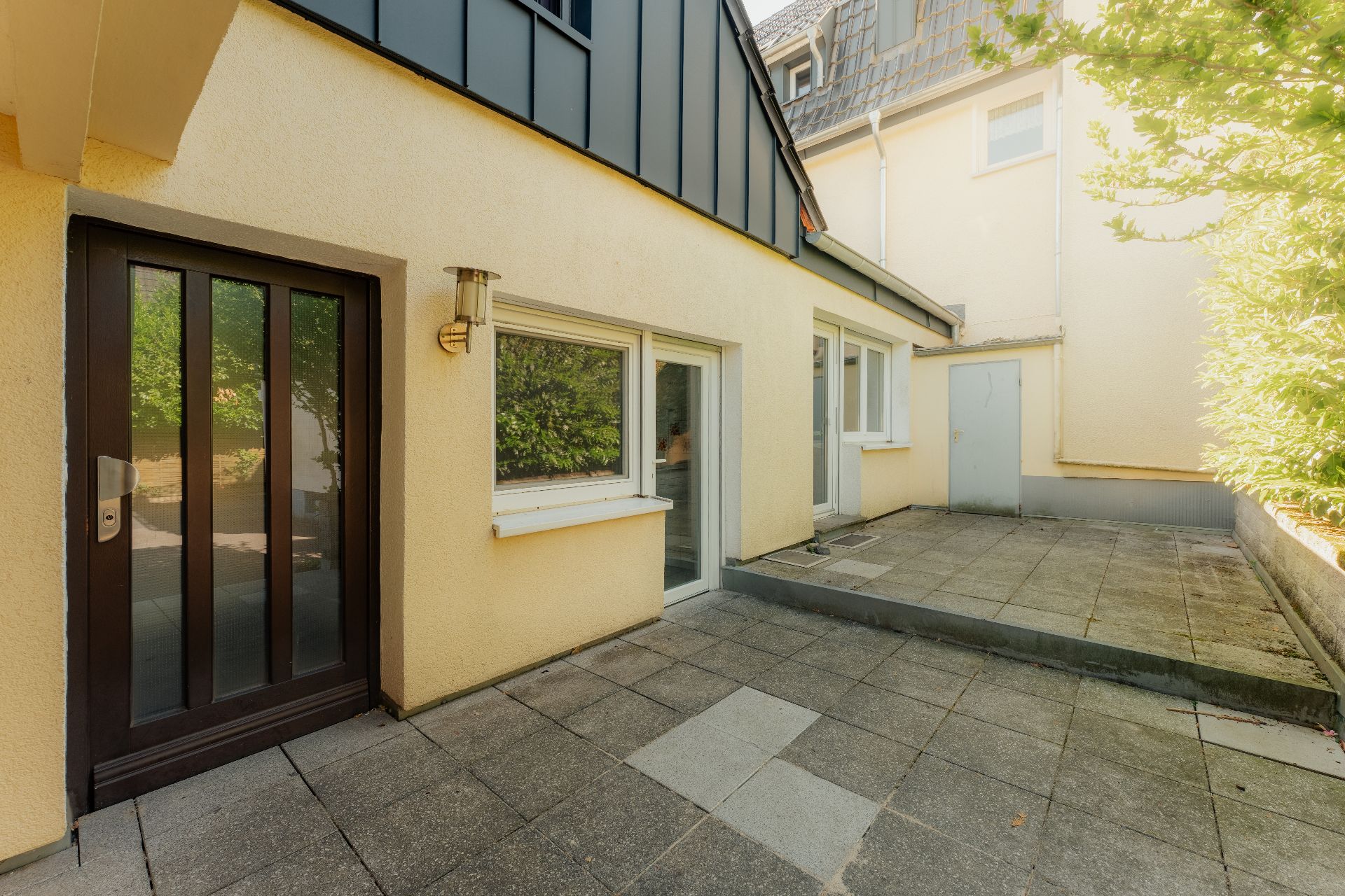 Gepflasterte Terrasse mit Glastür und Fenstern an einem gelben Wohnhaus, umgeben von Pflanzen.