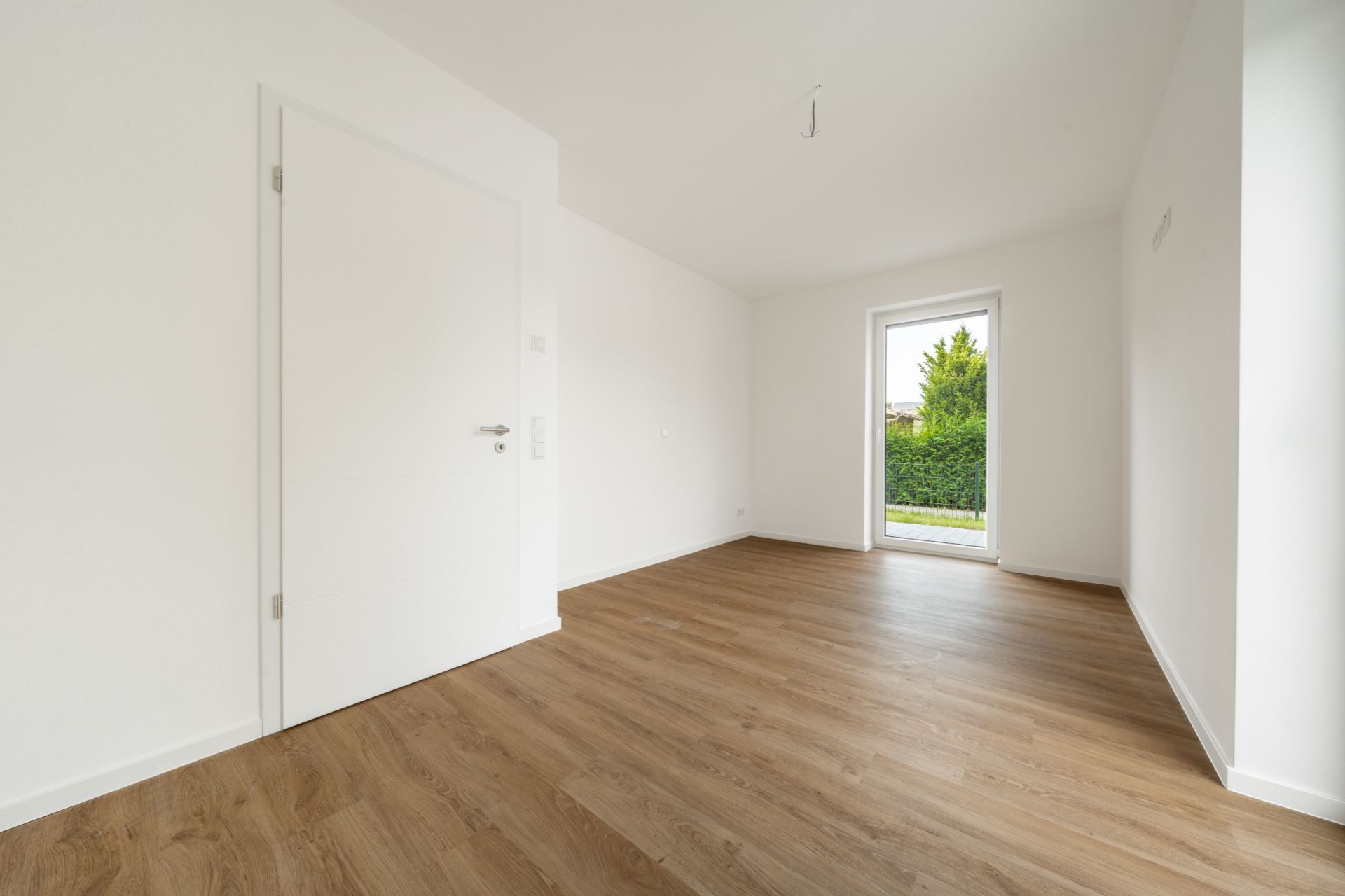Leerstehendes Zimmer mit Holzboden, weißer Tür und bodentiefem Fenster zum Garten.