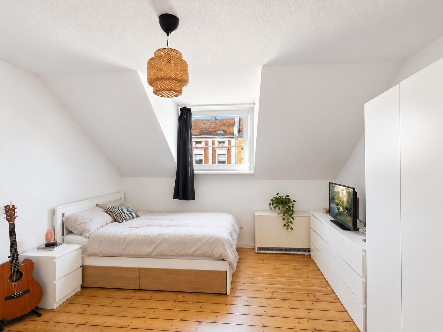 Helles Schlafzimmer mit Doppelbett, Schrank, Kommode, Gitarre und Fenster unter einer Dachschräge.