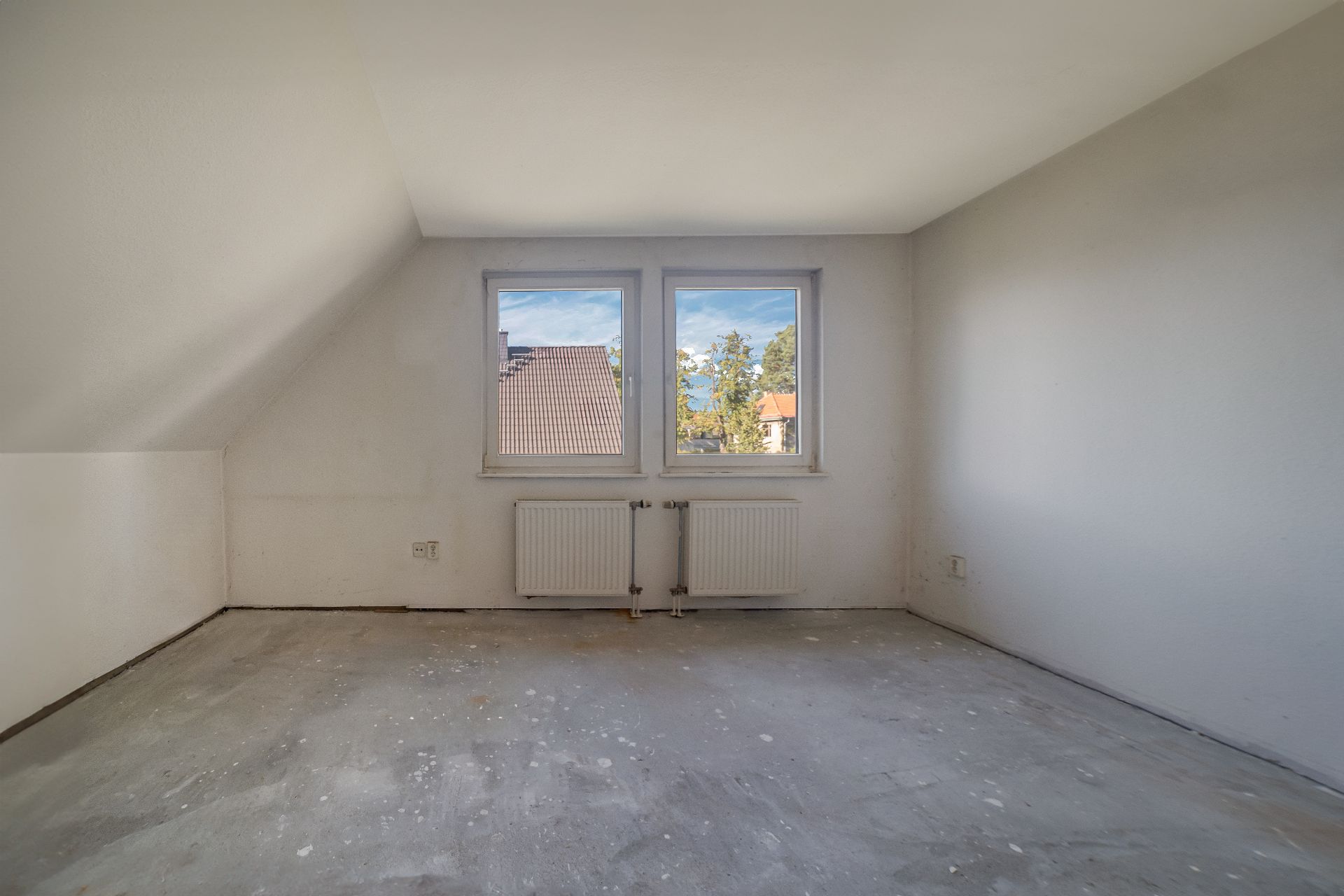 Leeres Zimmer mit zwei Fenstern, weiß gestrichenen Wänden und unrenoviertem Boden.