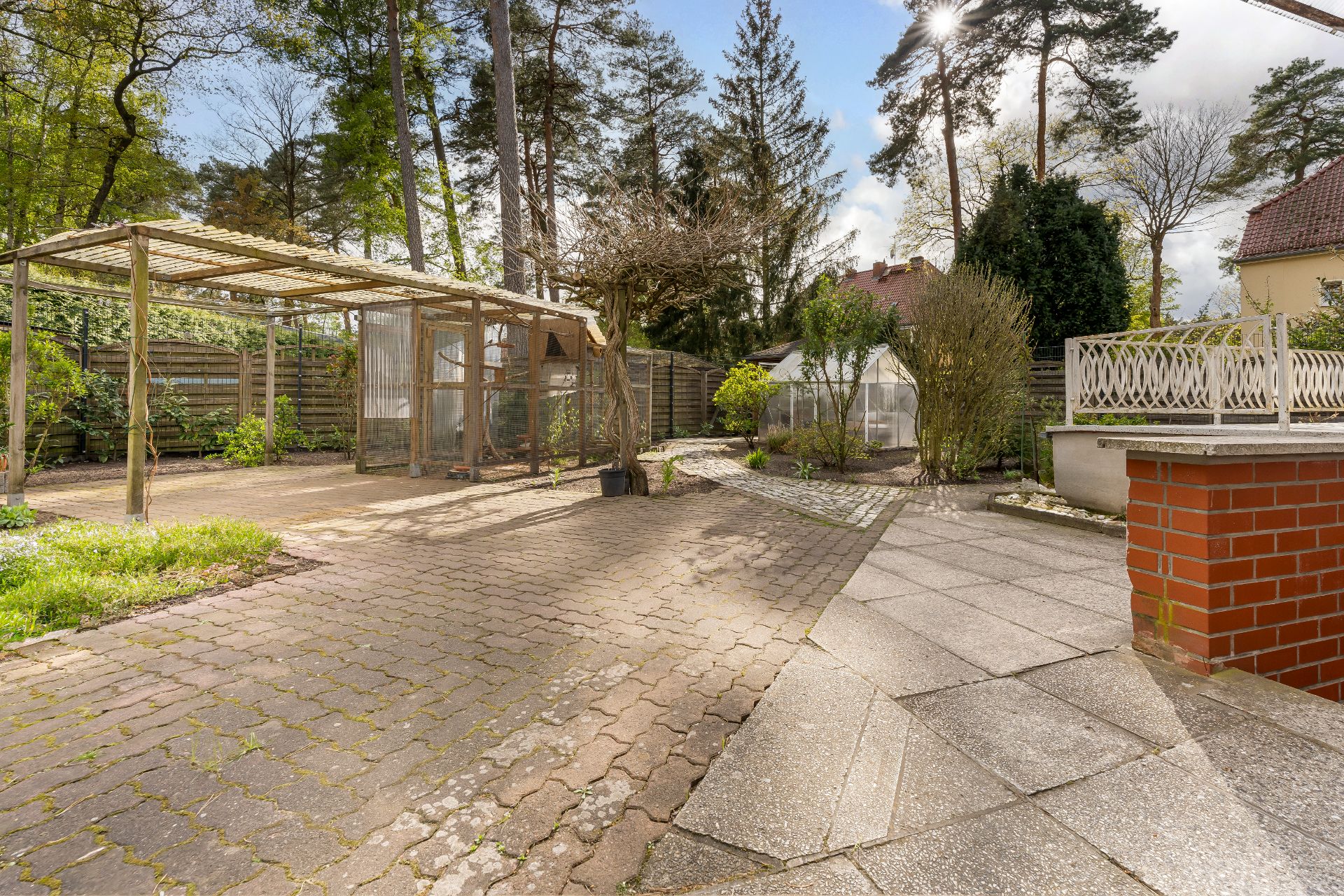 Gepflasterter Garten mit Holzüberdachung, Gewächshaus und Bäumen im Hintergrund.