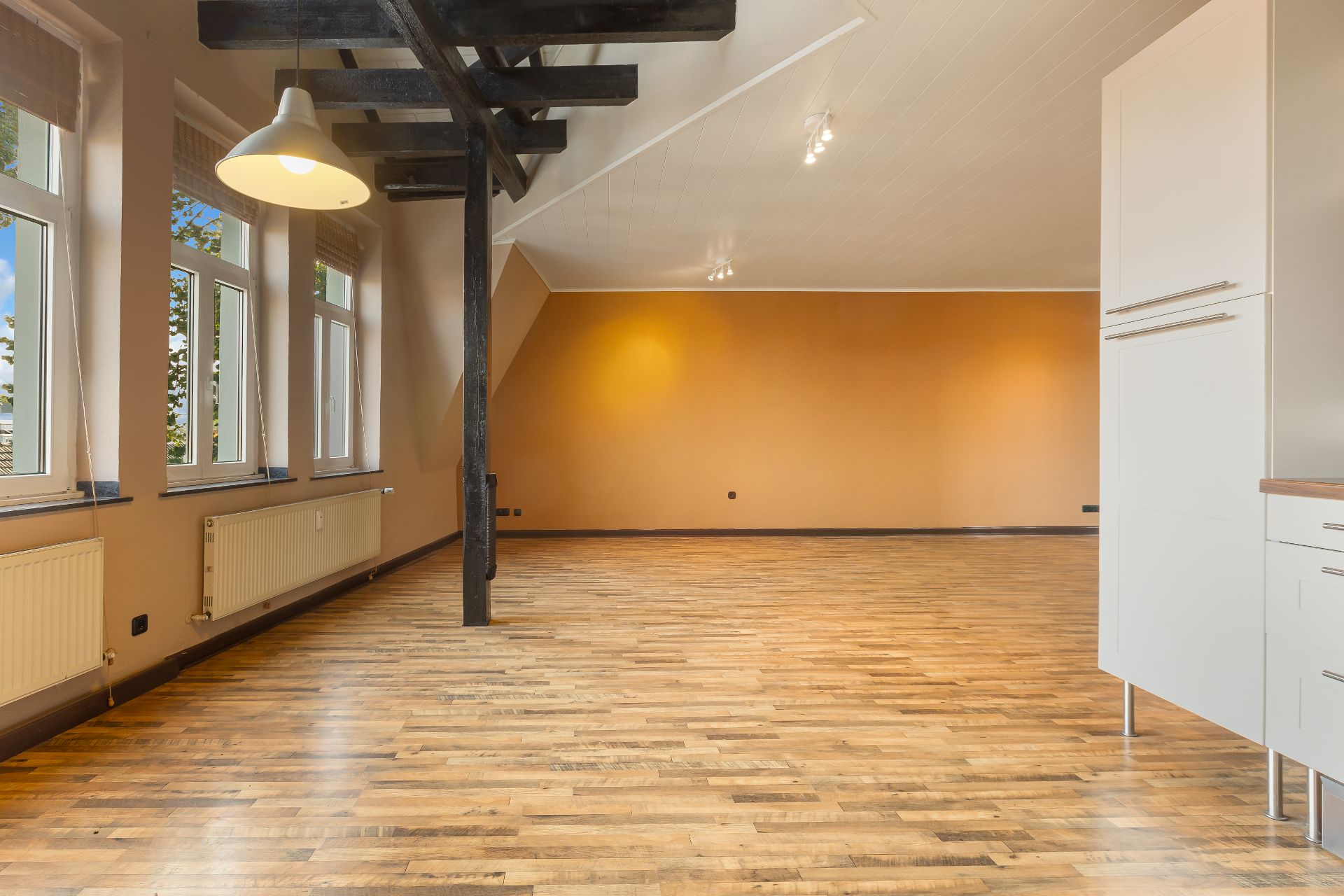Helles, leeres Zimmer mit Holzfußboden, großen Fenstern und sichtbaren Deckenbalken.
