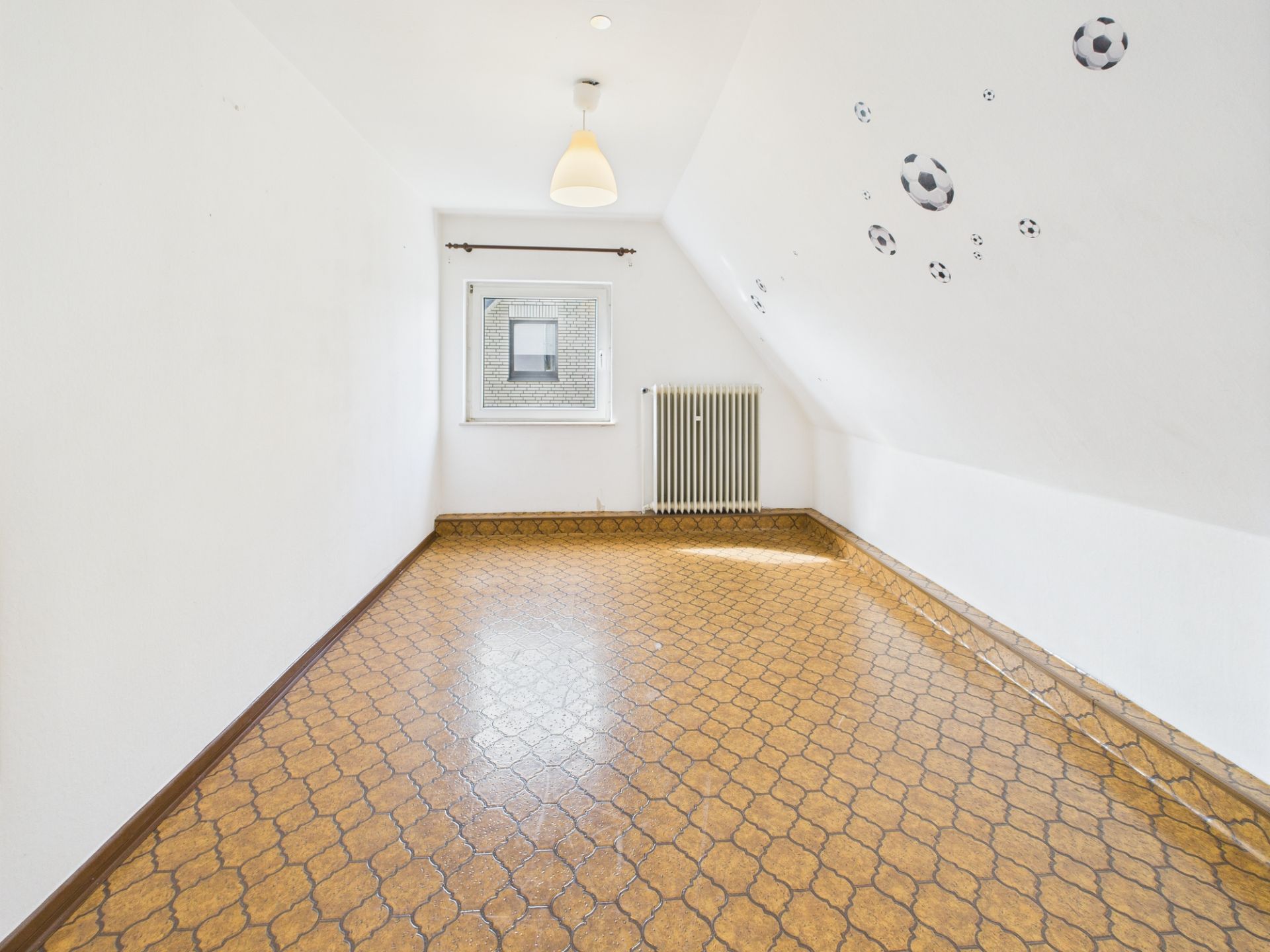 Leeres Zimmer mit braunem Boden, Fenster, Heizung und Wandaufklebern von Fußbällen.