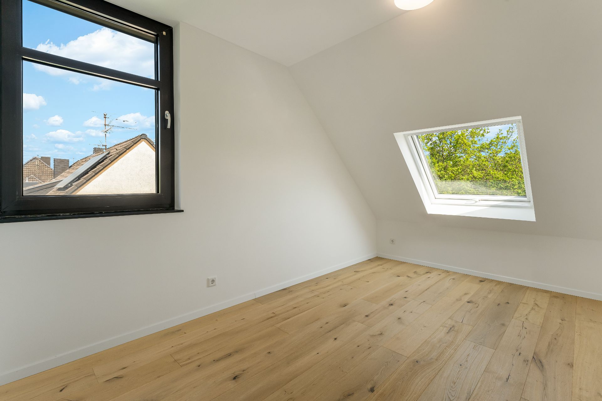 Heller Raum mit Holzfußboden, Dachschräge und zwei Fenstern, eines im Dach.