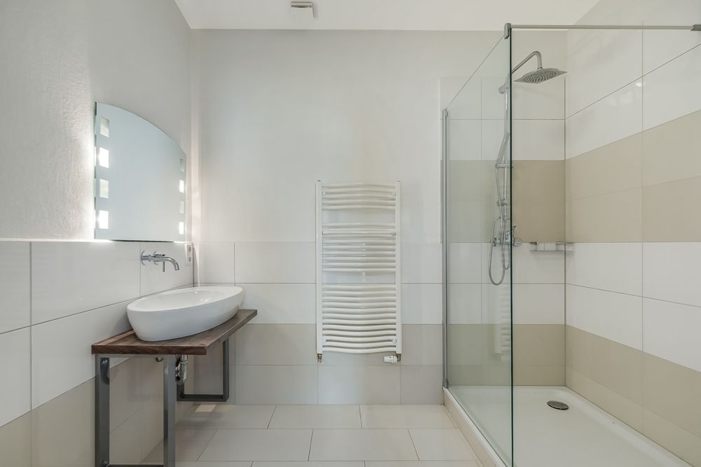 Modernes Badezimmer mit Waschbecken, beleuchtetem Spiegel, Handtuchheizkörper und Glasdusche.