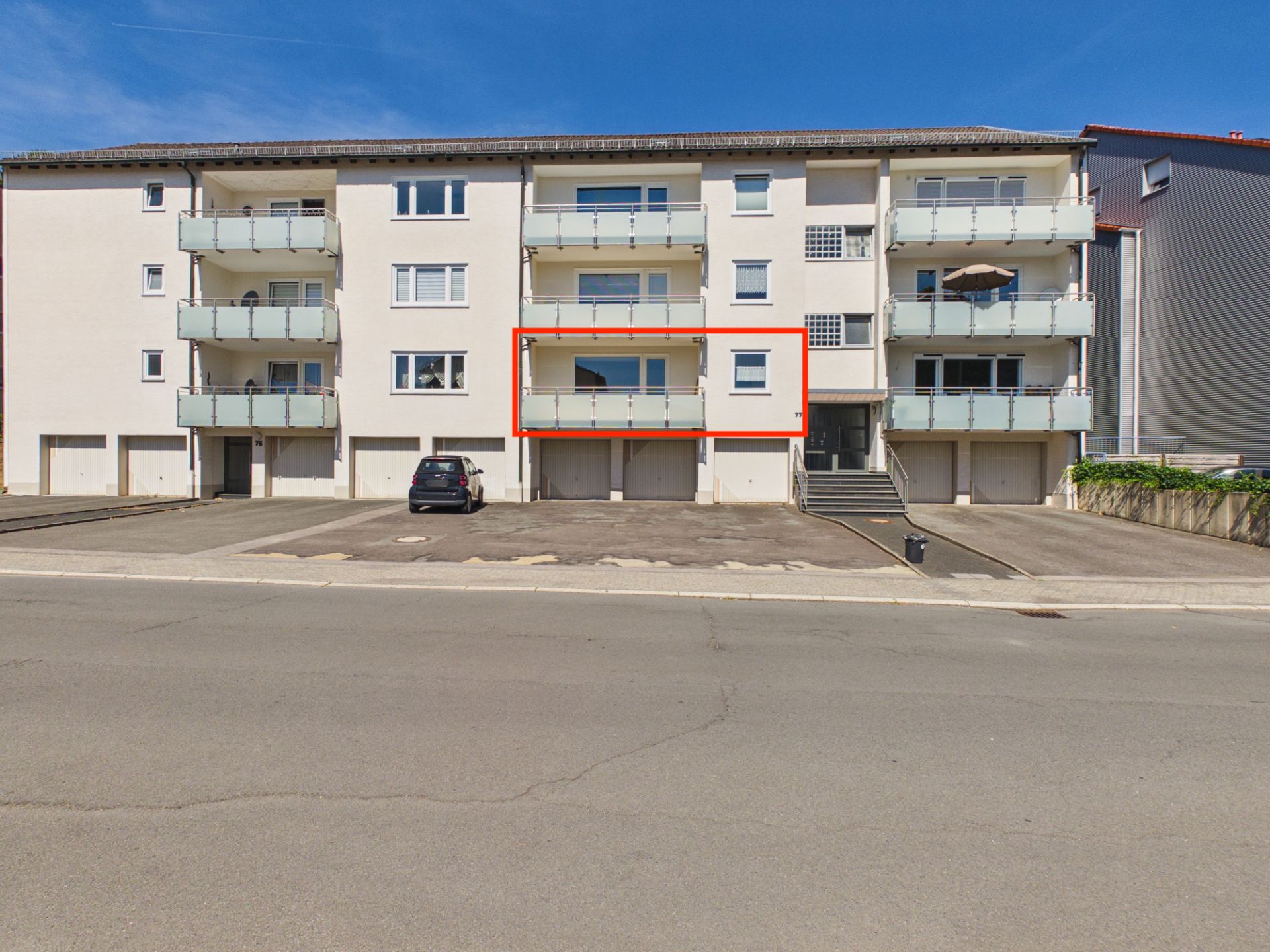 Mehrfamilienhaus mit markiertem Balkon und mehreren Parkplätzen vor dem Gebäude.