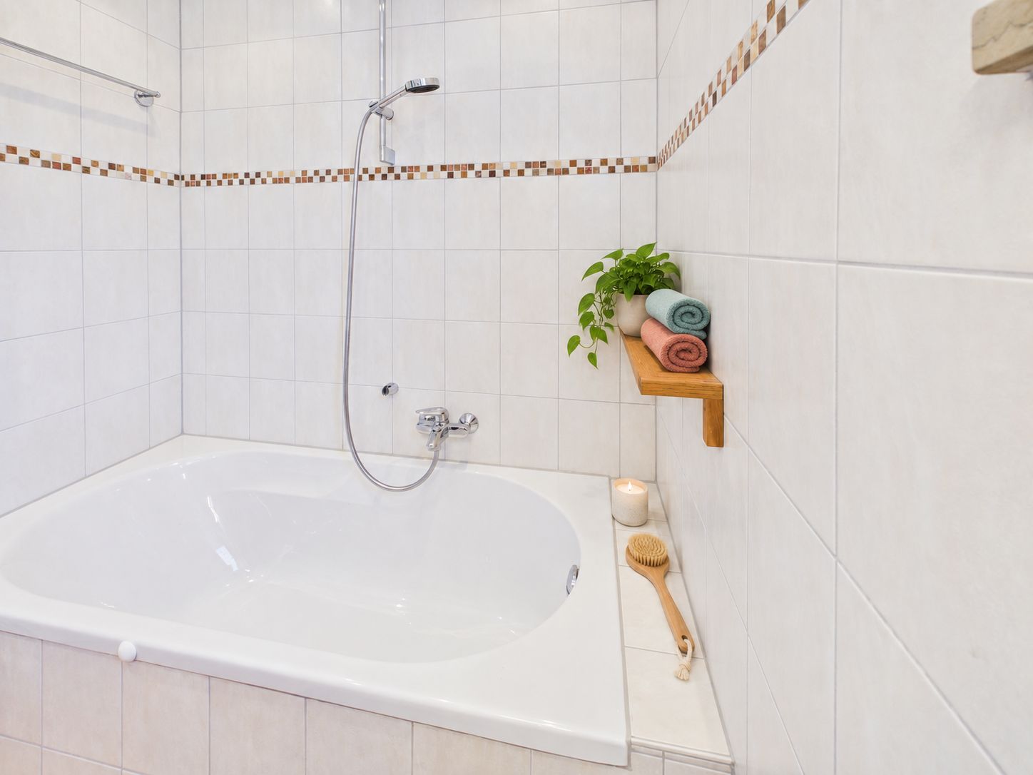 Bad mit weißer Badewanne, gefliesten Wänden, Duscheinrichtung und Regal mit Handtüchern und Pflanze.