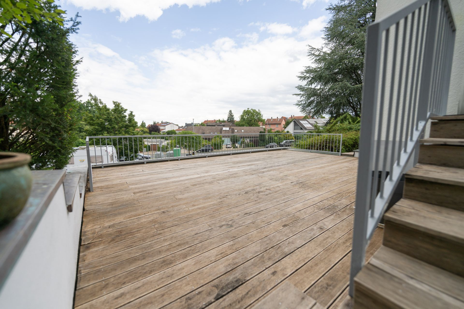 Große Holzterrasse mit Geländer und Treppe, Blick auf Nachbarhäuser und Bäume.