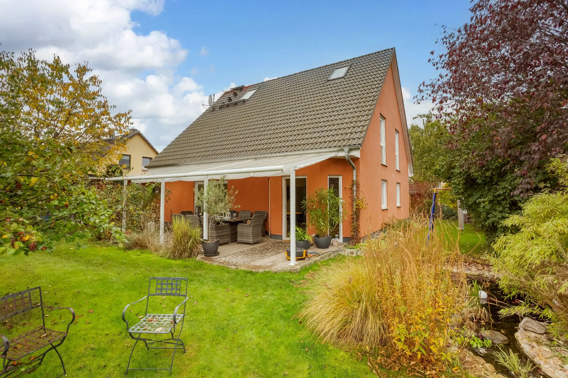 Zweistöckiges Haus mit Terrasse und Garten, umgeben von Bäumen und Sträuchern.