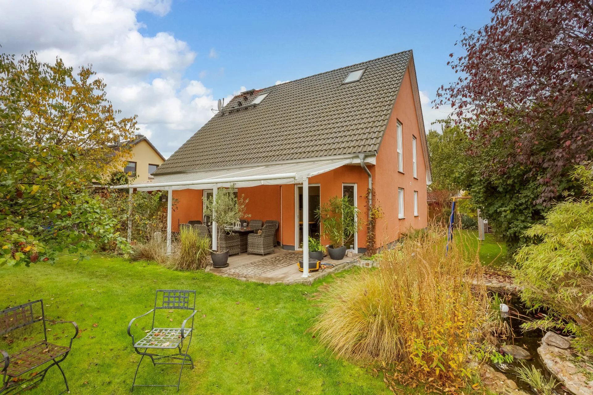 Zweistöckiges Haus mit Terrasse und Garten, umgeben von Bäumen und Sträuchern.
