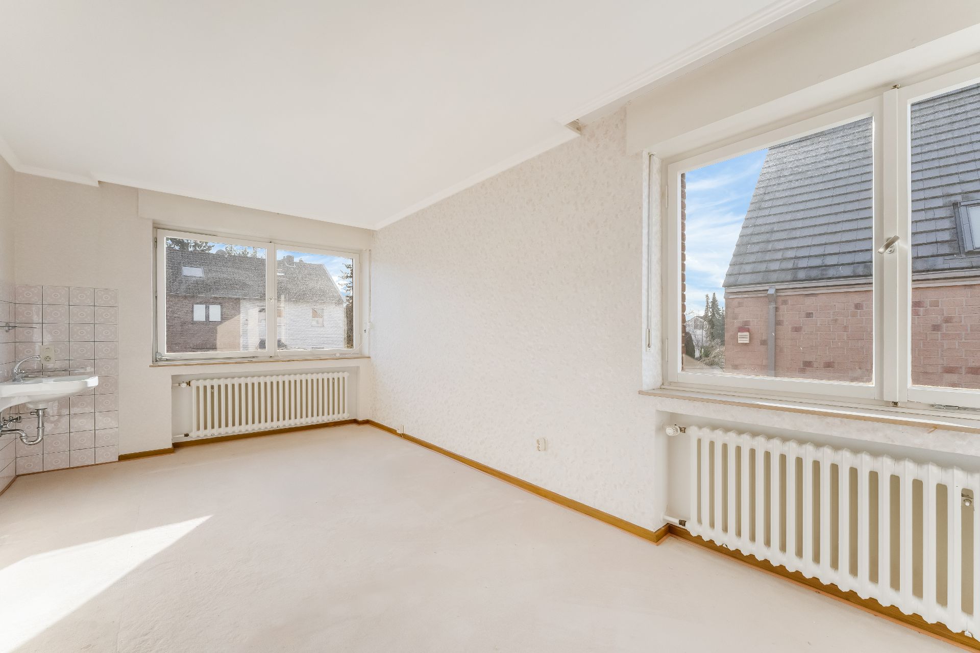 Leerstehendes, helles Zimmer mit zwei großen Fenstern, weißem Heizkörper und kleinem Waschbecken.