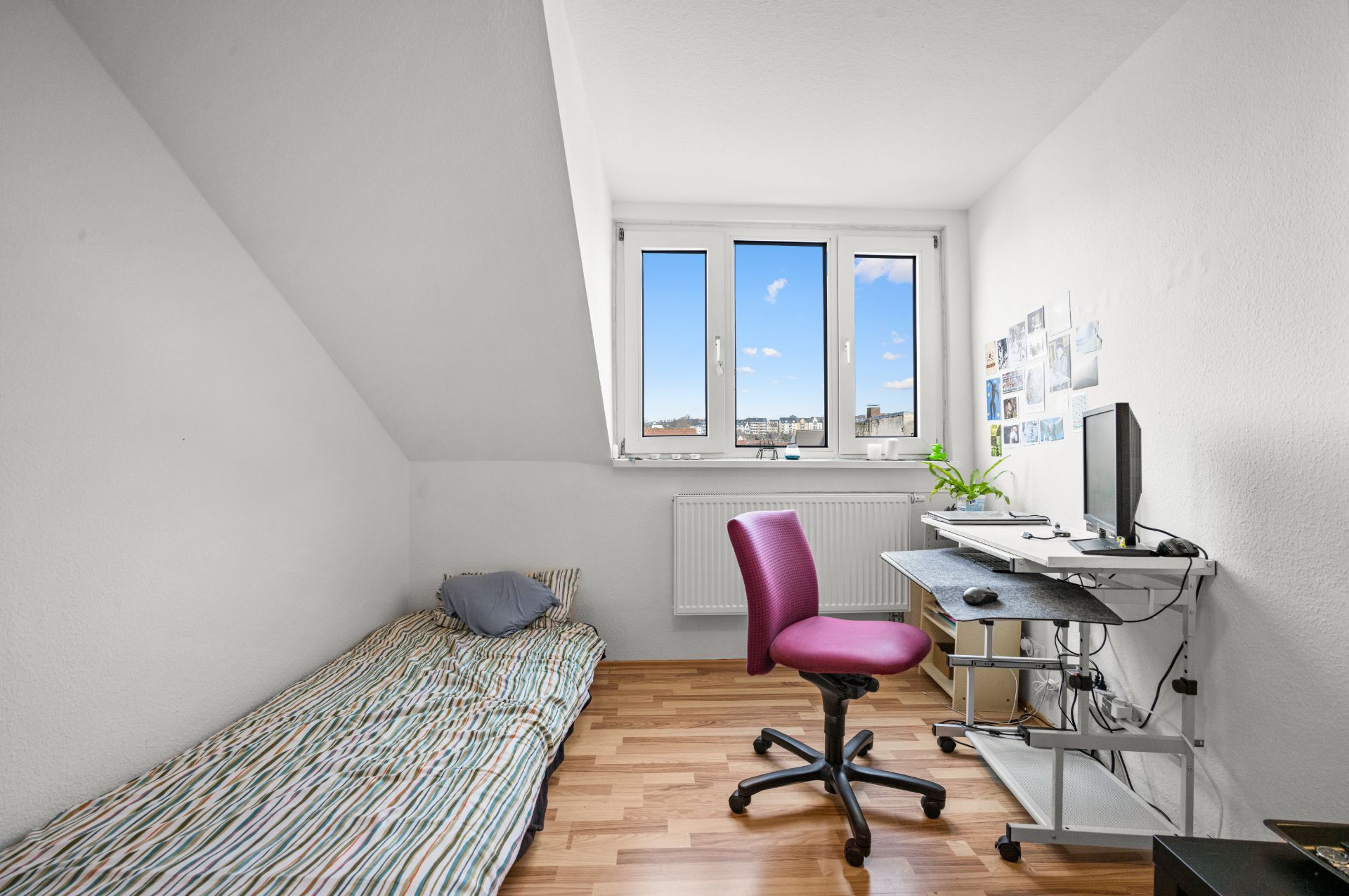 Kleines Zimmer mit Einzelbett, Schreibtisch, Stuhl und Fenster, auf hellem Holzfußboden.