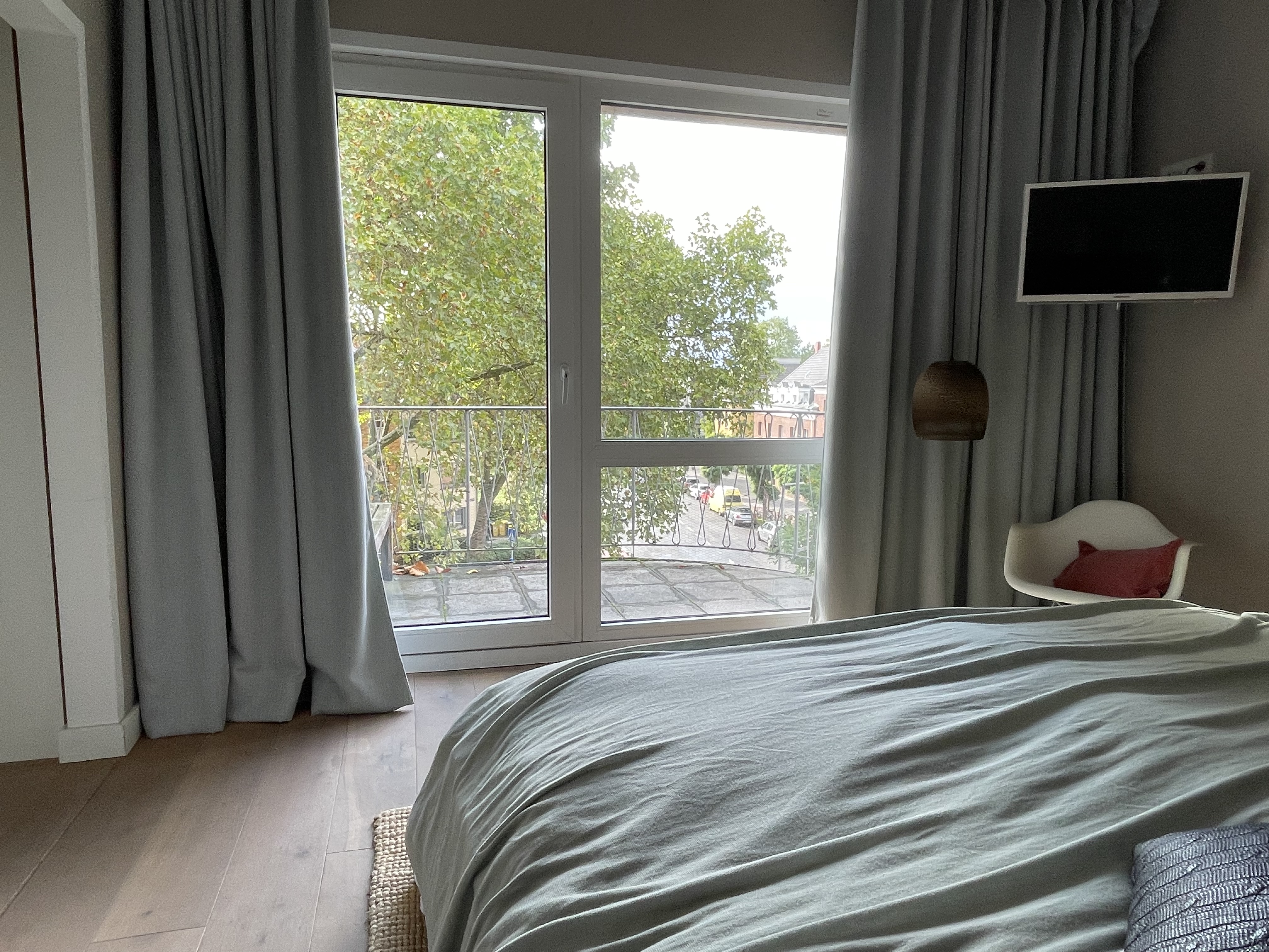 Schlafzimmer mit großem Fenster, Balkon, Stuhl und Fernseher, grüne Bäume draußen sichtbar.