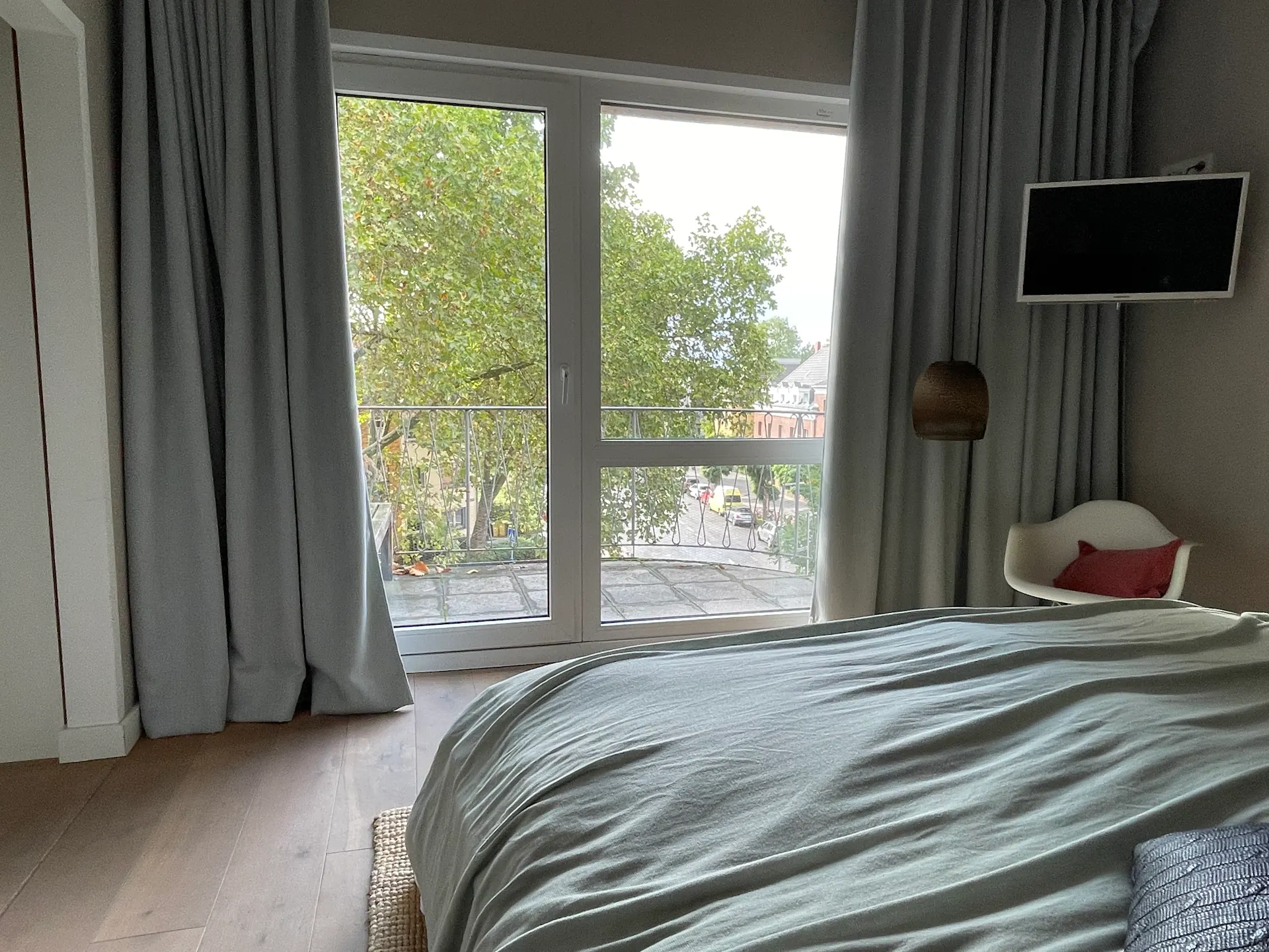 Schlafzimmer mit großem Fenster, Balkon, Stuhl und Fernseher, grüne Bäume draußen sichtbar.