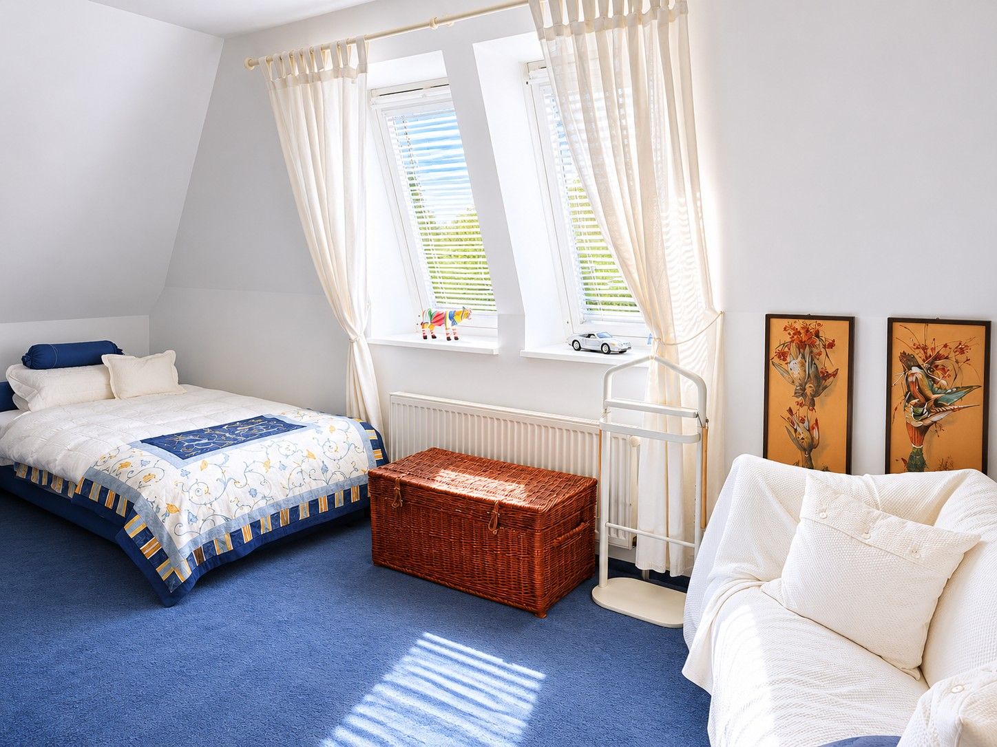 Helles Schlafzimmer mit Einzelbett, weißem Sofa, Korbtruhe und zwei Fenstern mit Vorhängen.