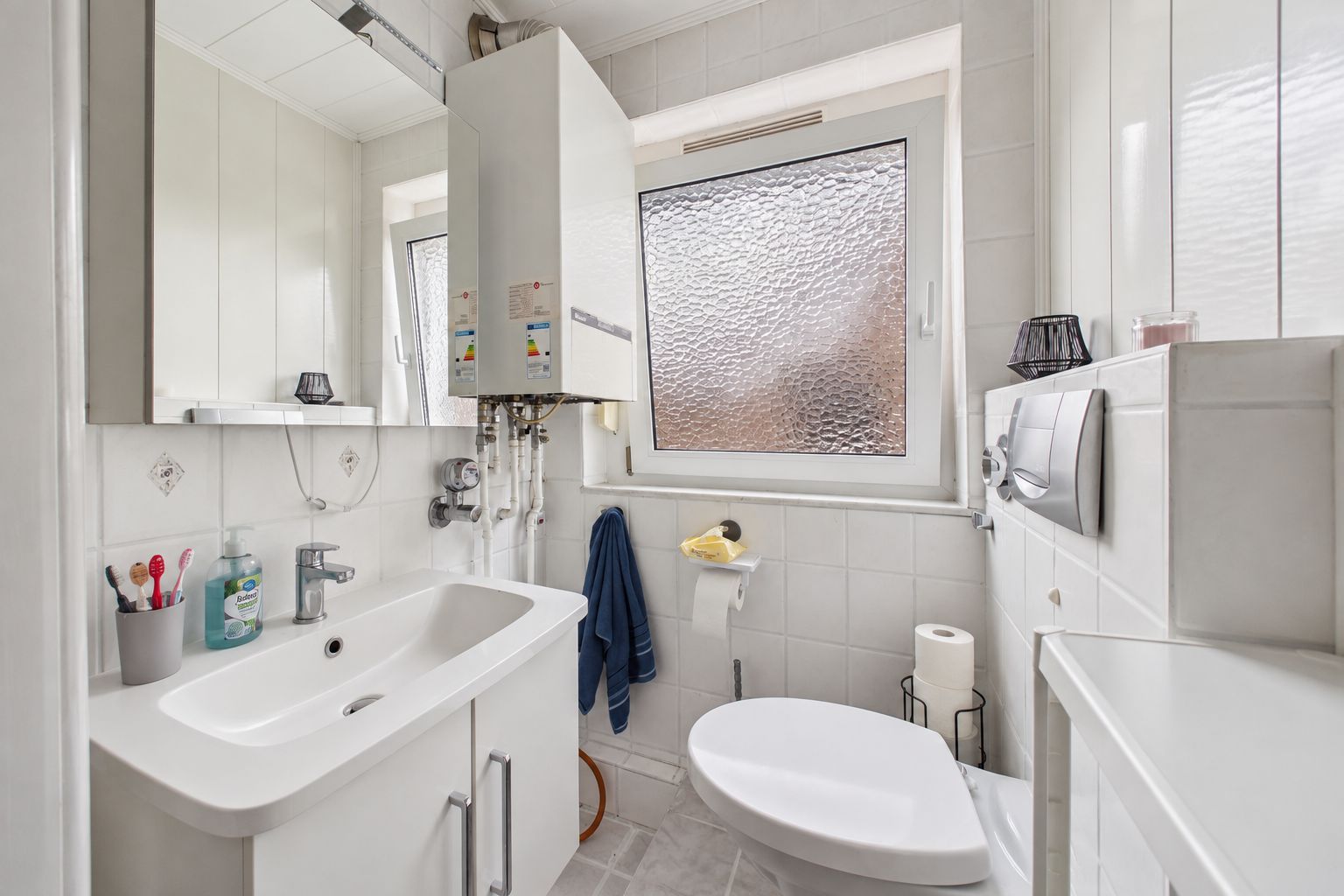 Kleines, helles Badezimmer mit Waschbecken, Toilette, Spiegel und Fenster mit Milchglas.
