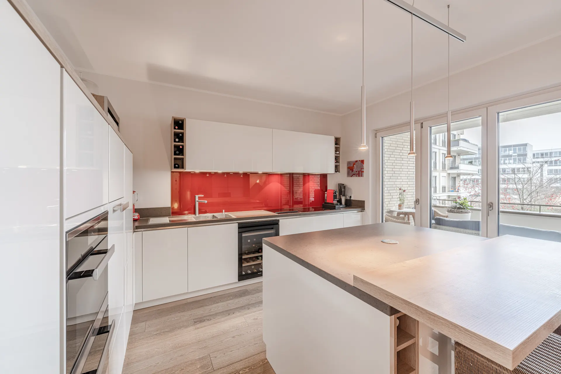Moderne Küche mit weißen Schränken, roter Rückwand und großem Küchenblock, Zugang zum Balkon.