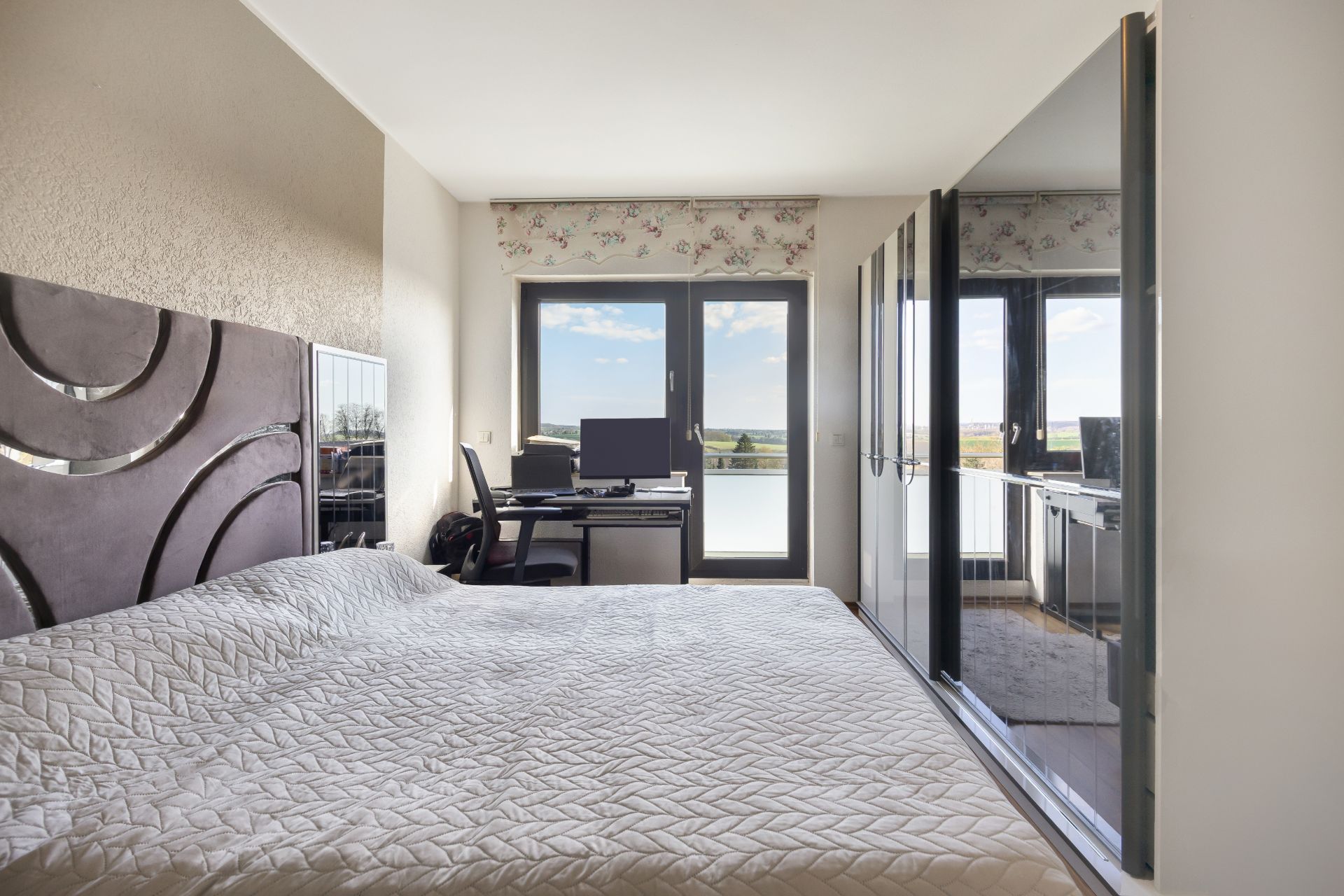 Schlafzimmer mit Bett, Schreibtisch, Stuhl und großem Fenster mit Blick ins Freie.