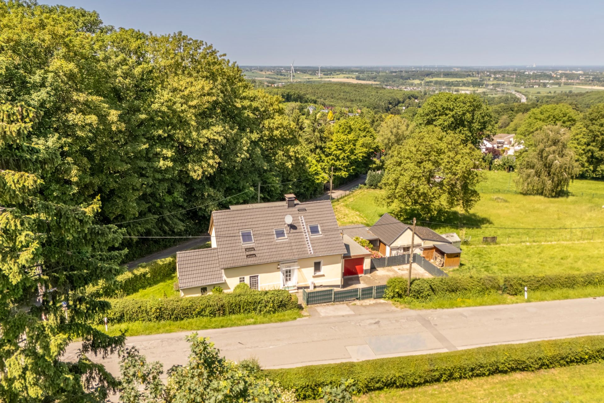 Haus mit grauem Dach und Garten an einer Landstraße, von Bäumen und Wiese umgeben.