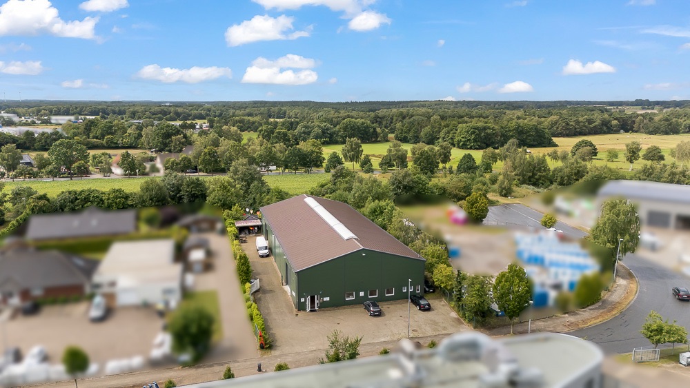 Grünes Gebäude mit Parkplatz und Bäumen, im Hintergrund Felder und Wald.