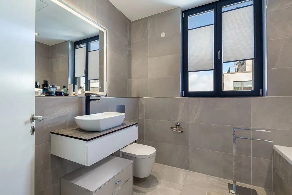 Modernes Badezimmer mit großem Spiegel, Waschbecken, WC und zwei Fenstern mit Jalousien.