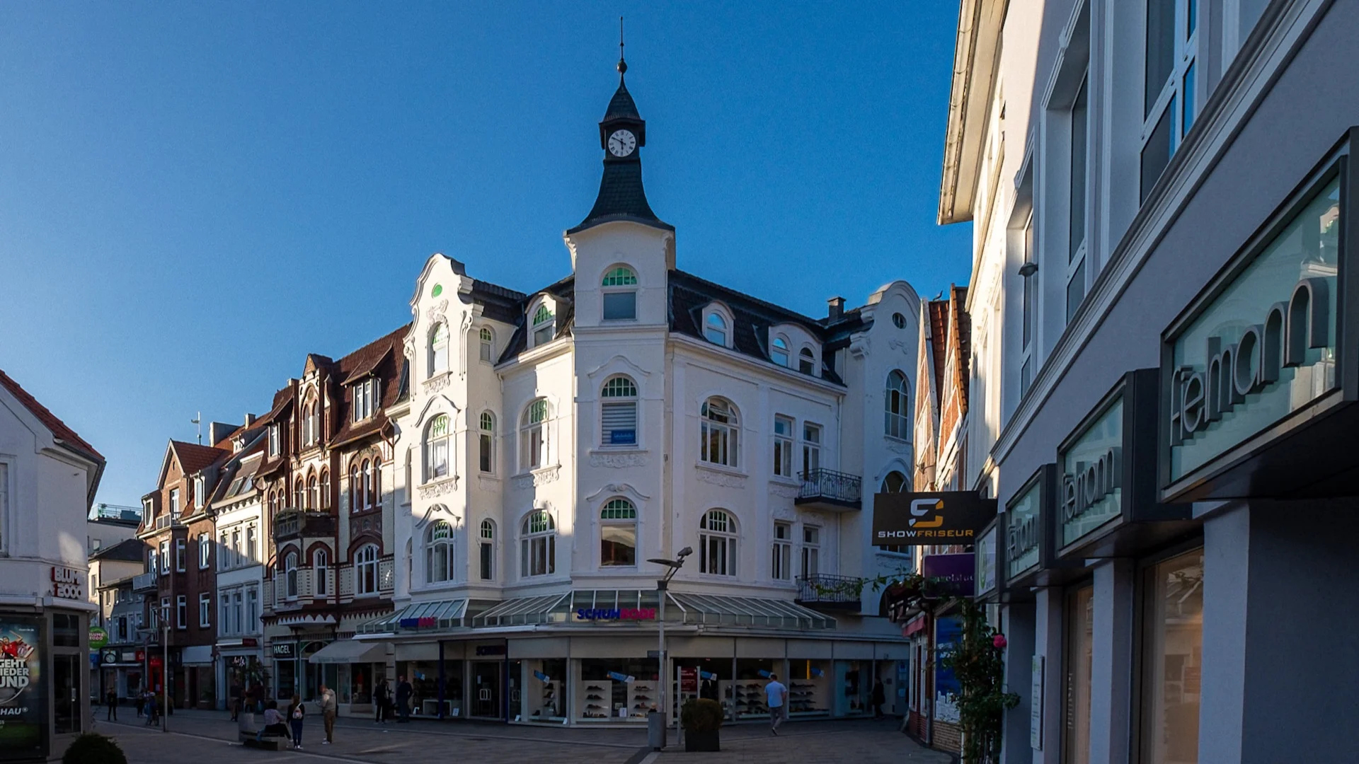 Weiße Wohnhausfassade mit Uhrturm und Geschäften im Erdgeschoss an einer Einkaufsstraße.