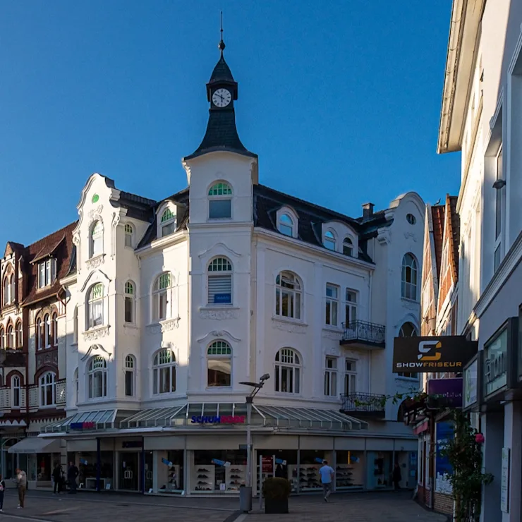 Weiße Wohnhausfassade mit Uhrturm und Geschäften im Erdgeschoss an einer Einkaufsstraße.