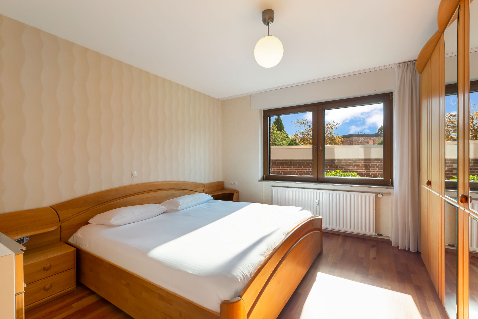 Helles Schlafzimmer mit Doppelbett, Holzschränken und großem Fenster mit Blick nach draußen.