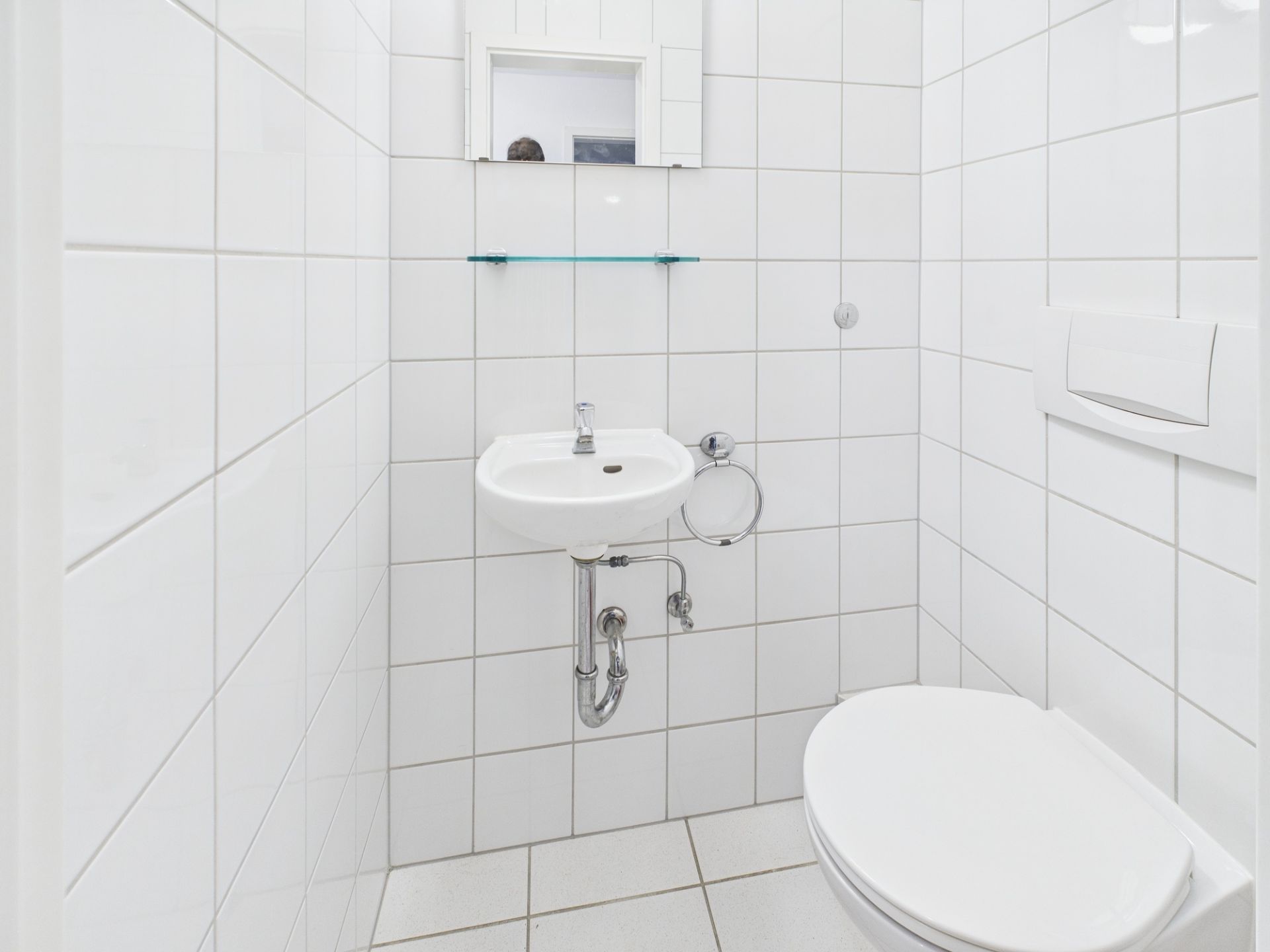 Kleines Badezimmer mit weiß gefliesten Wänden, Waschbecken, Toilette und Spiegel.