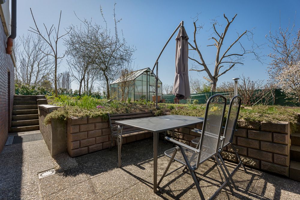Terrasse mit Tisch, zwei Stühlen, Sonnenschirm und Blick auf ein Gewächshaus im Garten.