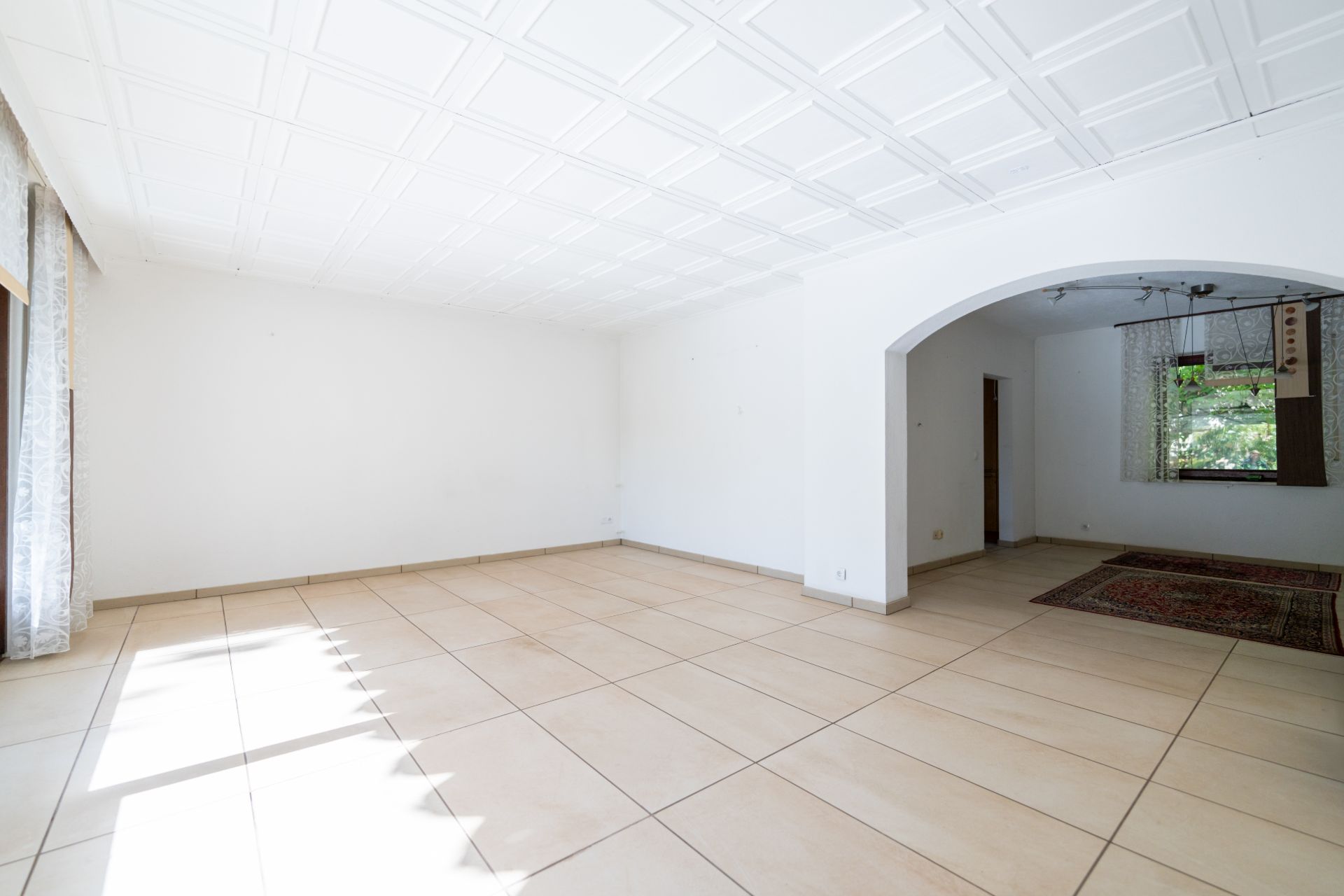 Heller, leerer Raum mit Fliesenboden, weiße Wände, Fenster und ein Durchgang zu weiterem Zimmer.