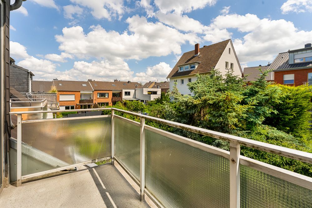 Balkon mit Geländer, Blick auf Nachbarhäuser und grüne Bäume unter blauem Himmel mit Wolken.