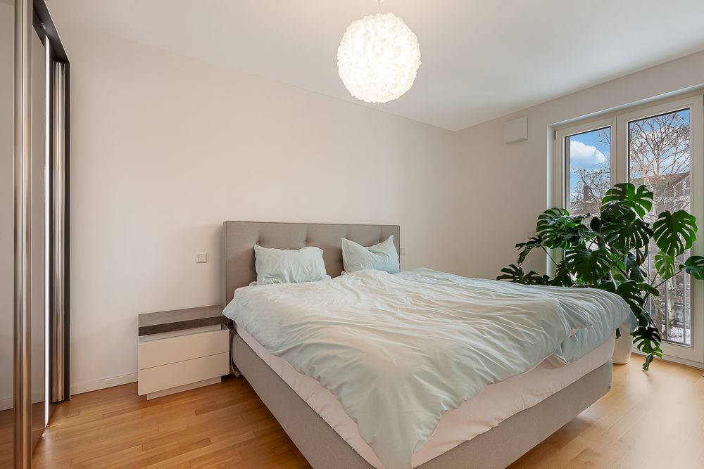 Helles Schlafzimmer mit Doppelbett, Nachttisch, großem Fenster und grüner Zimmerpflanze.