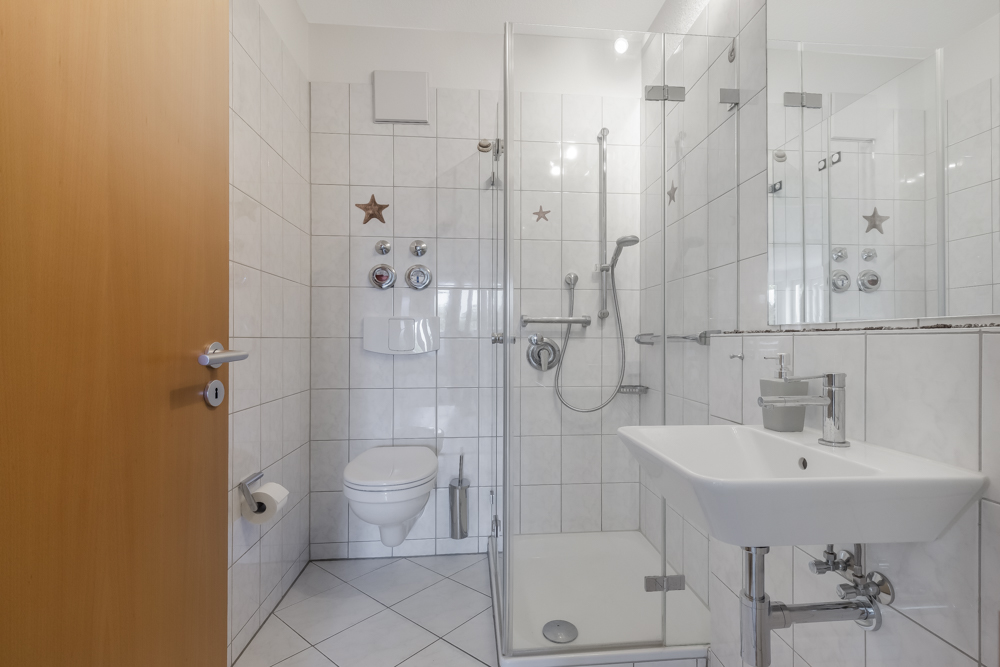 Modernes Badezimmer mit Dusche, Waschbecken und Wand-WC, weiße Fliesen an Wänden und Boden.