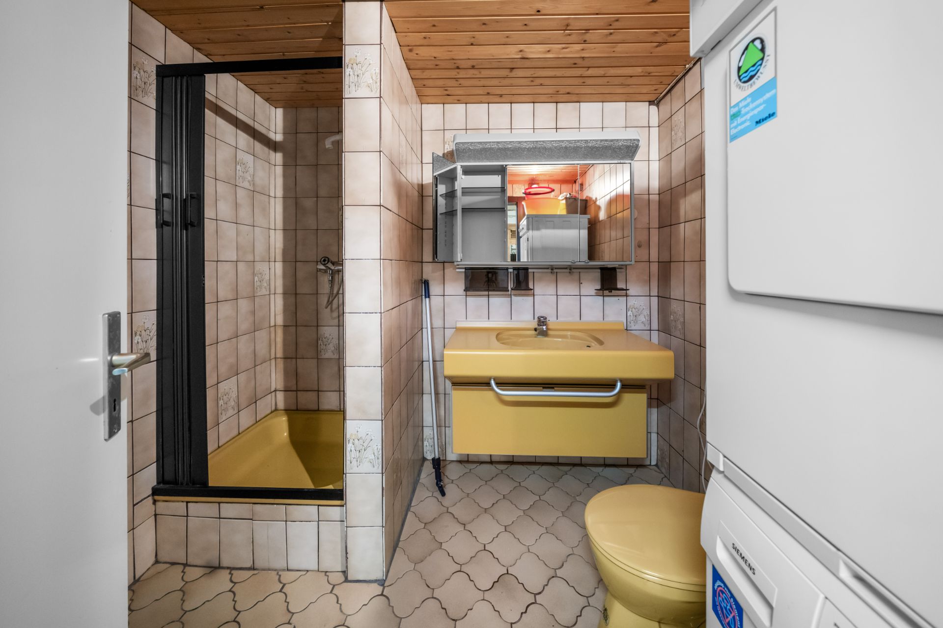 Badezimmer mit gelbem Waschbecken, WC, Dusche und Waschmaschine, Holzdecke und Fliesenwänden.