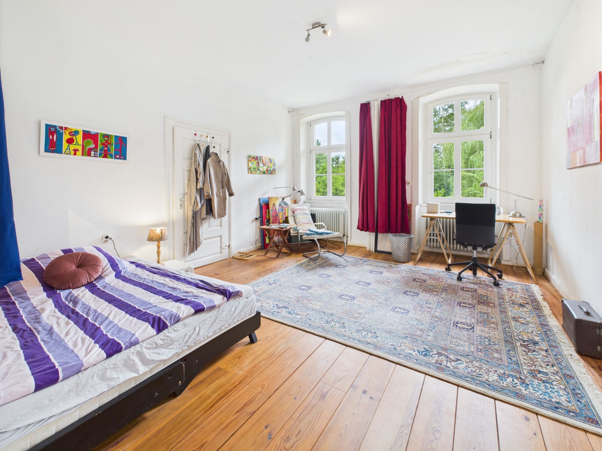 Helles Schlafzimmer mit Bett, Schreibtisch, großem Teppich und zwei Fenstern mit roten Vorhängen.