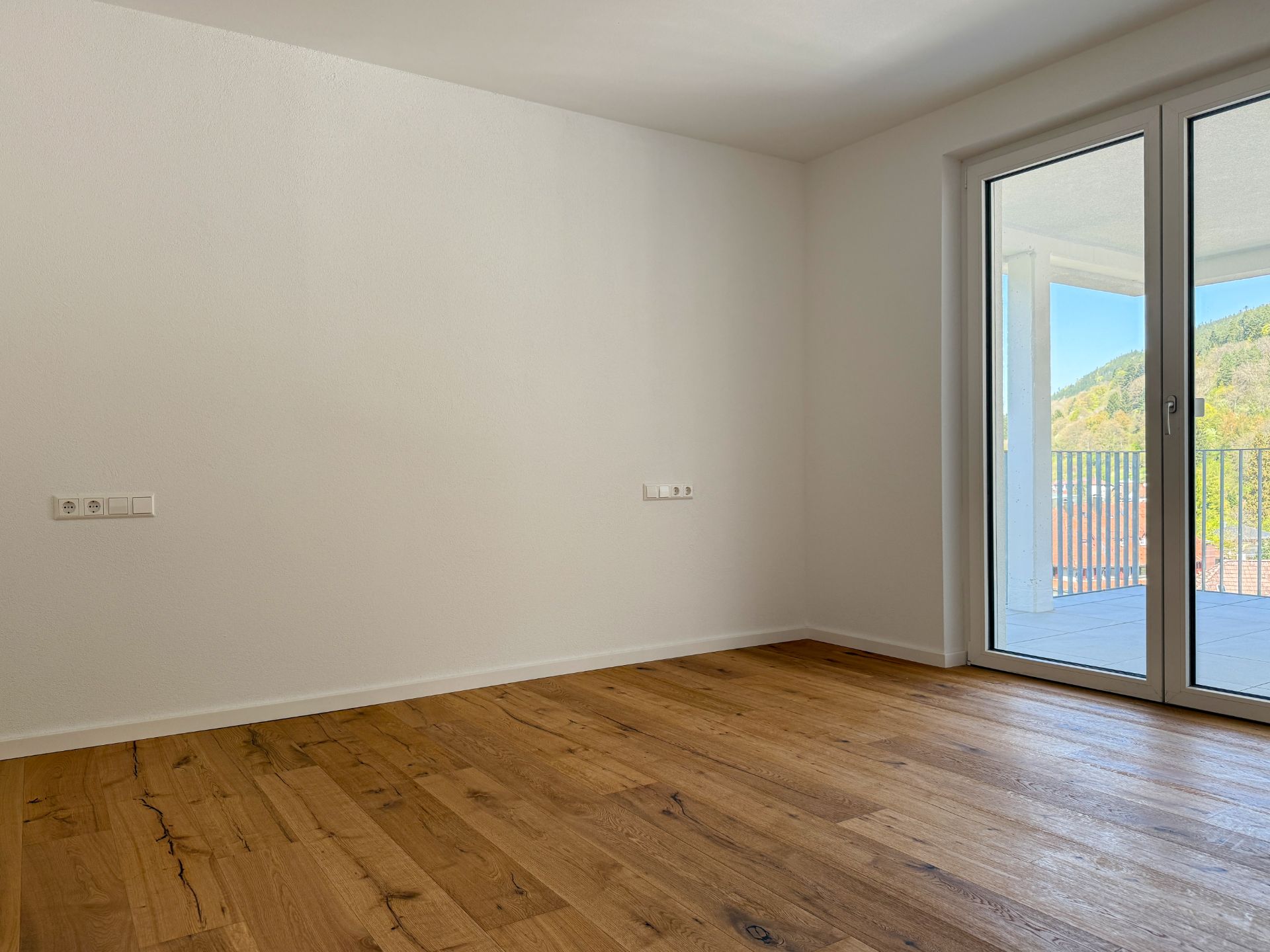 Leerer Raum mit Holzfußboden und großer Glastür zu einem Balkon mit Ausblick auf grüne Hügel.