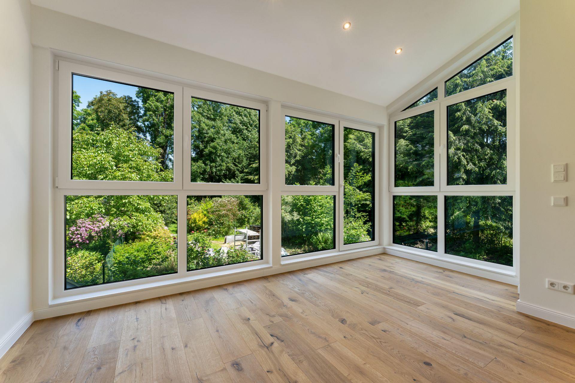 Heller Raum mit großen Fenstern und Blick auf einen grünen Garten, Holzboden und weiße Wände.