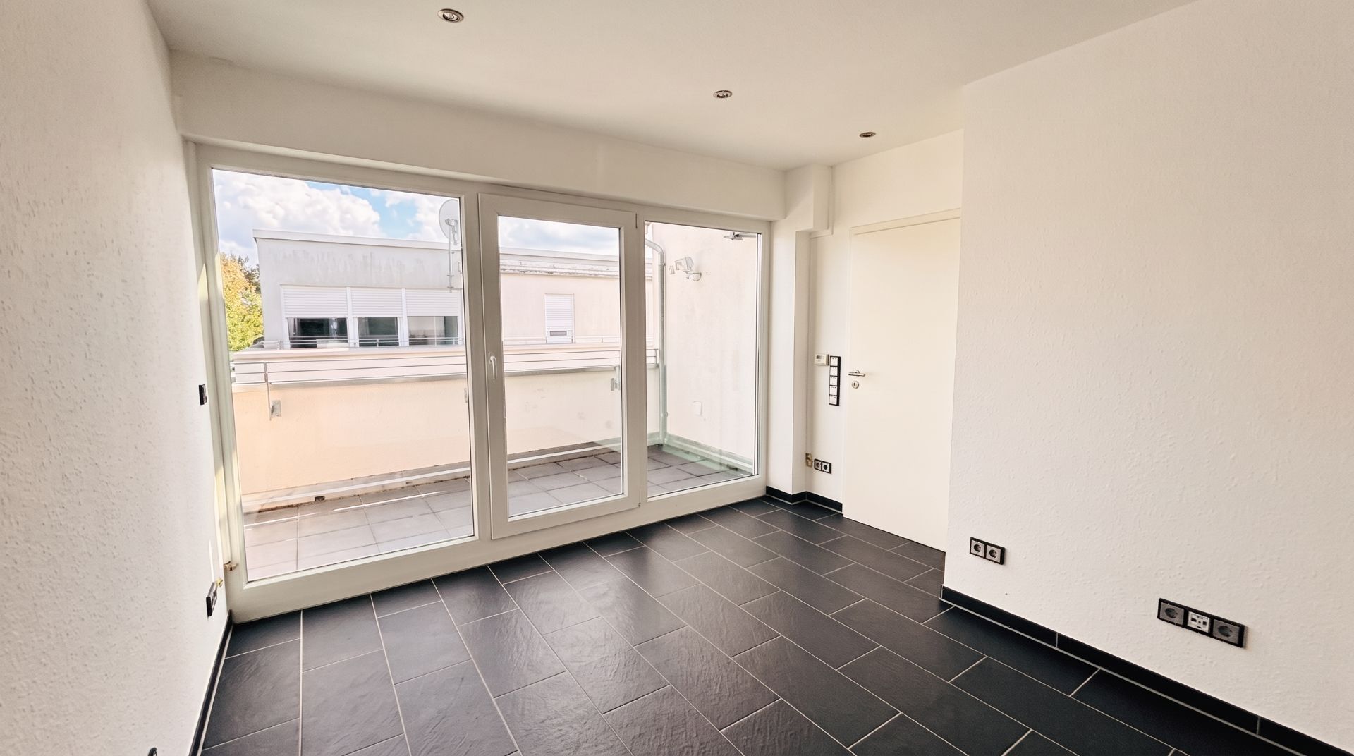 Heller Raum mit schwarzen Fliesen, großen Fenstern und Zugang zu einem Balkon.